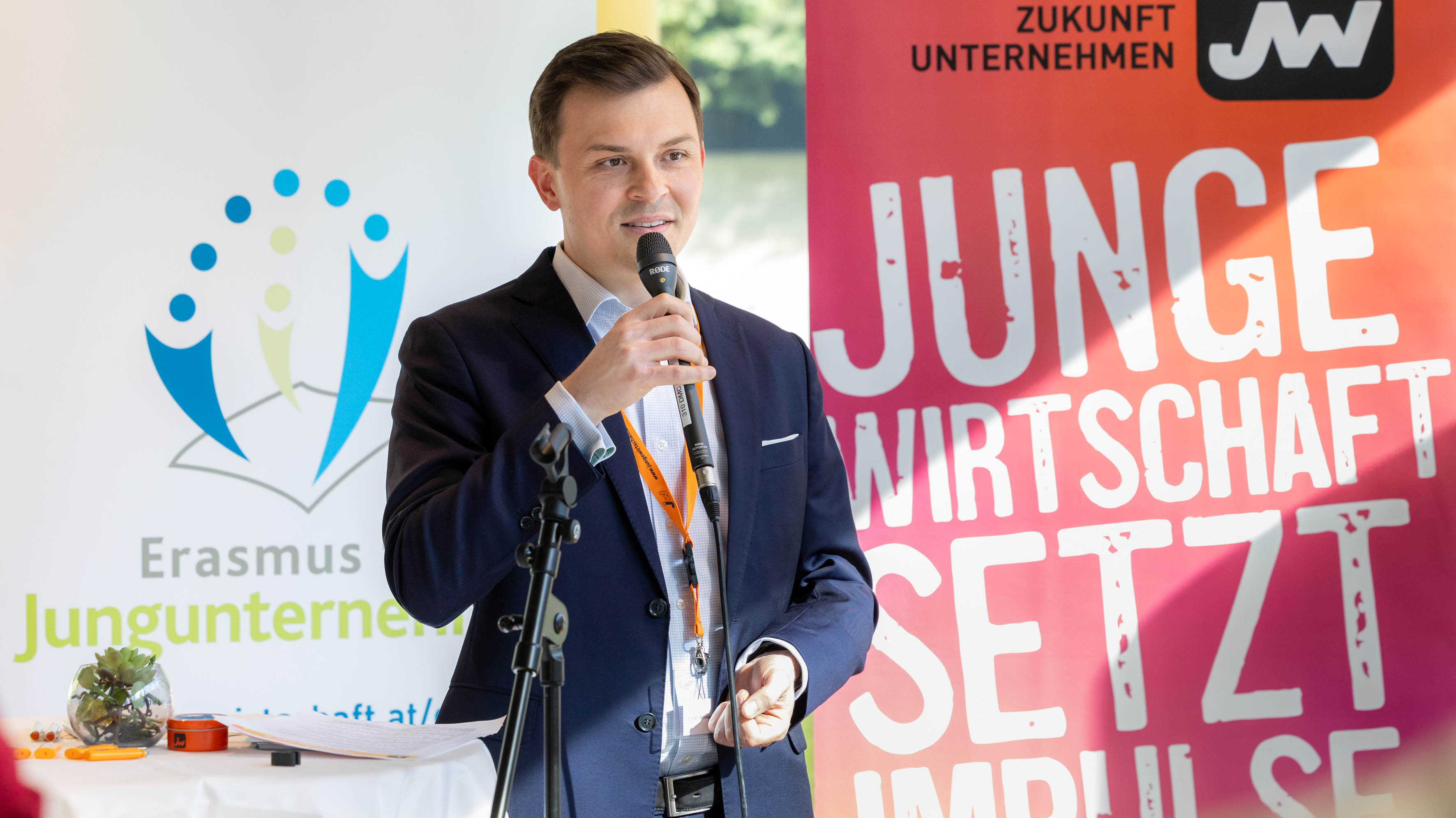Eine Person mit Mikrofon in der rechten Hand steht auf einer B&uuml;hne. Im Hintergrund ist ein Roll-Up mit dem Schriftzug Junge Wirtschaft setzt Impulse und ein Plakat mit dem Schriftzug Erasmus Jungunternehmer