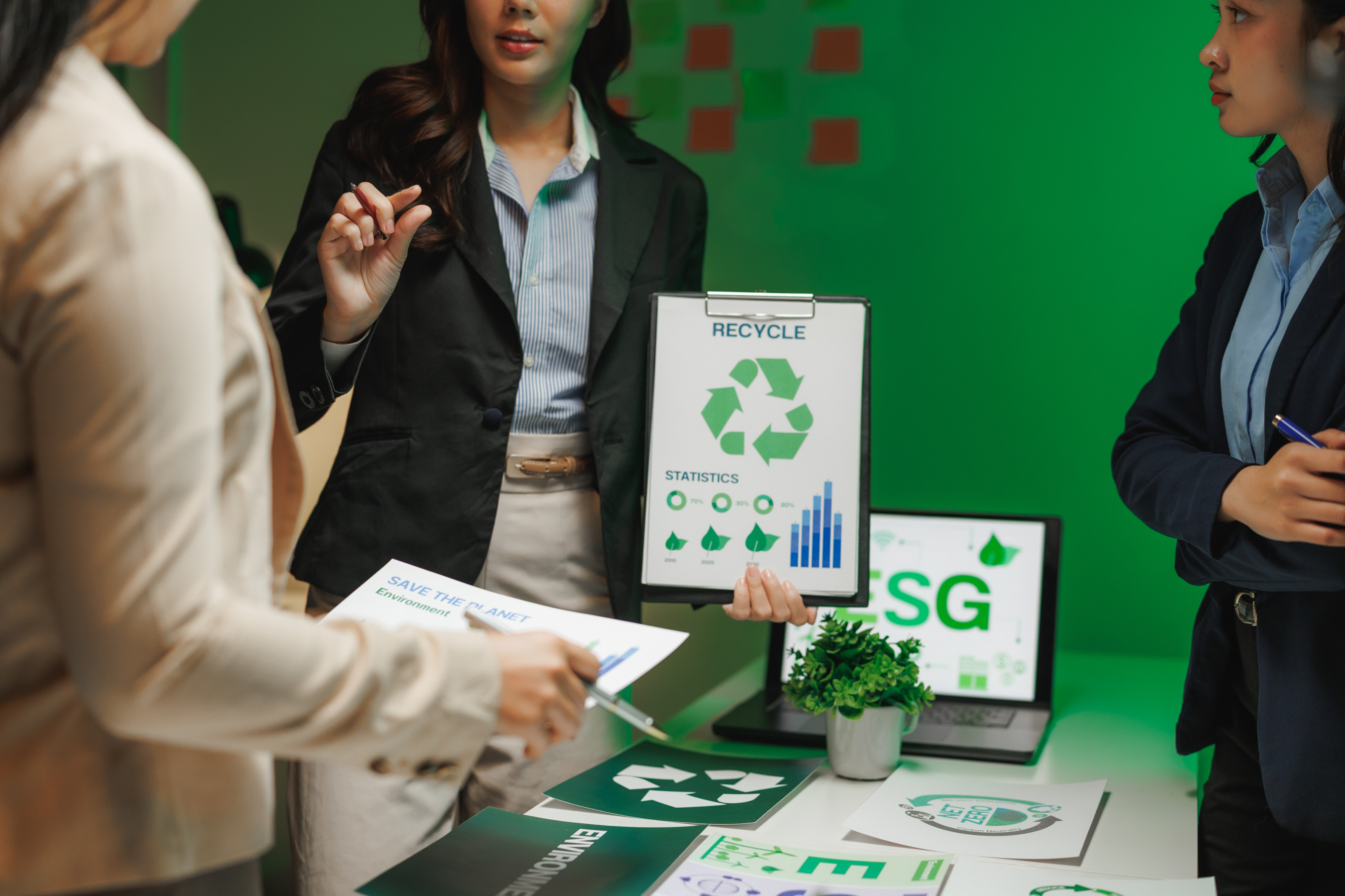 Um einen Tisch stehen drei Personen in Sakkos. Eine der Personen h&auml;lt ein Klemmbrett, auf dem ein Zettel mit einem Recyclingsymbol ist. Darunter sind Diagramme. Die Person, die das Klemmbrett h&auml;lt, spricht