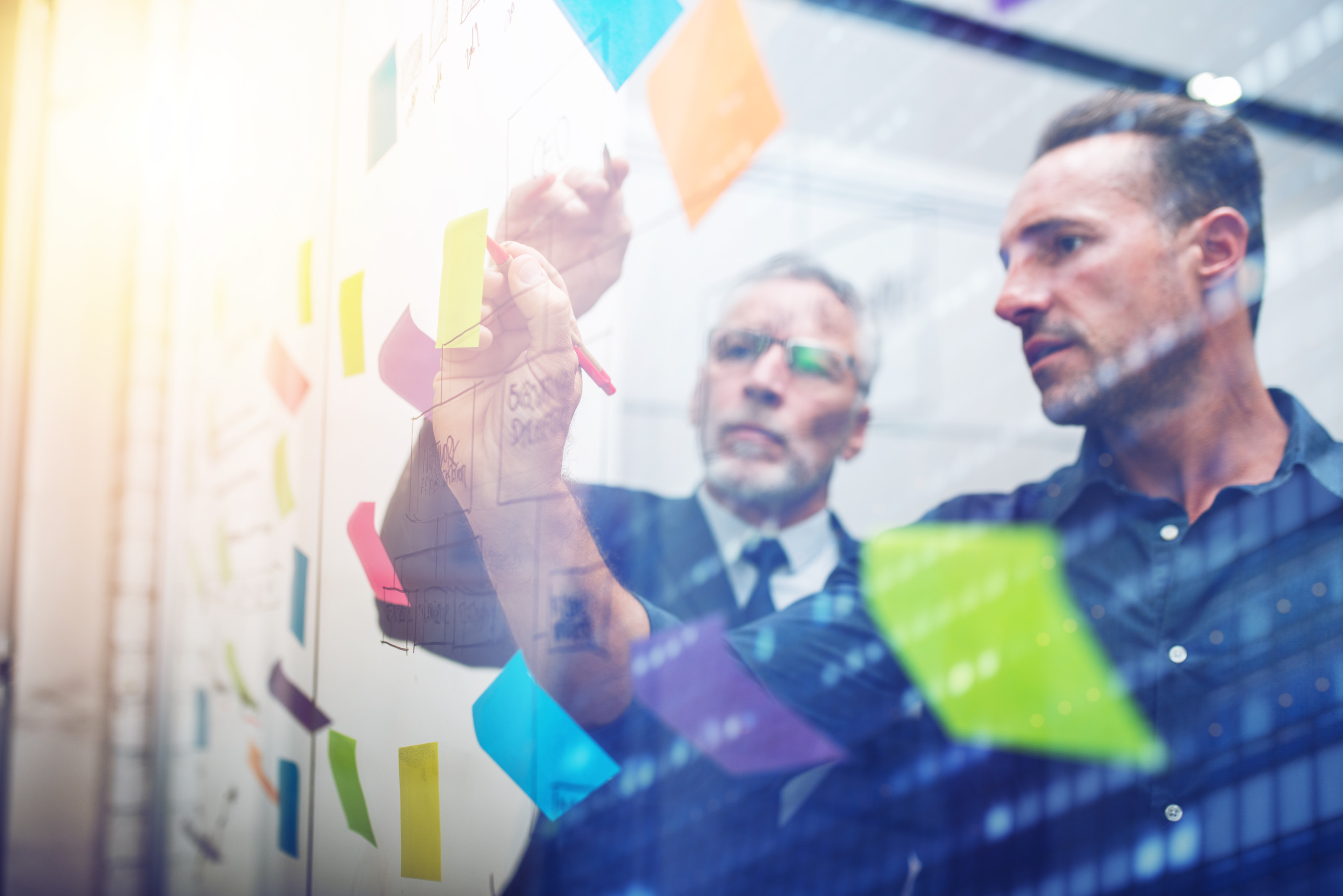 Personen in Businesskleidung stehen bei einem gl&auml;sernen Whiteboard und