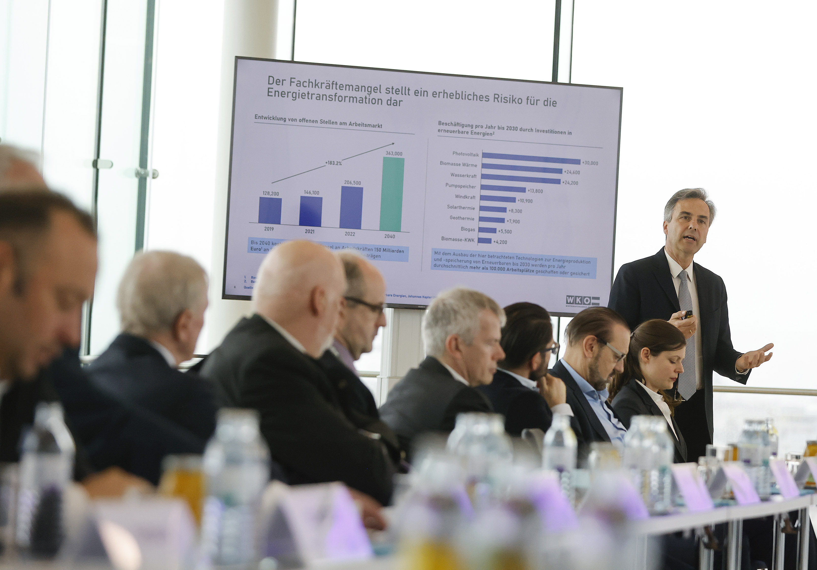 Stehende Person spricht zu anderen sitzende Personen vor einem Monitor mit Grafik zu Fachkr&auml;ftemangel