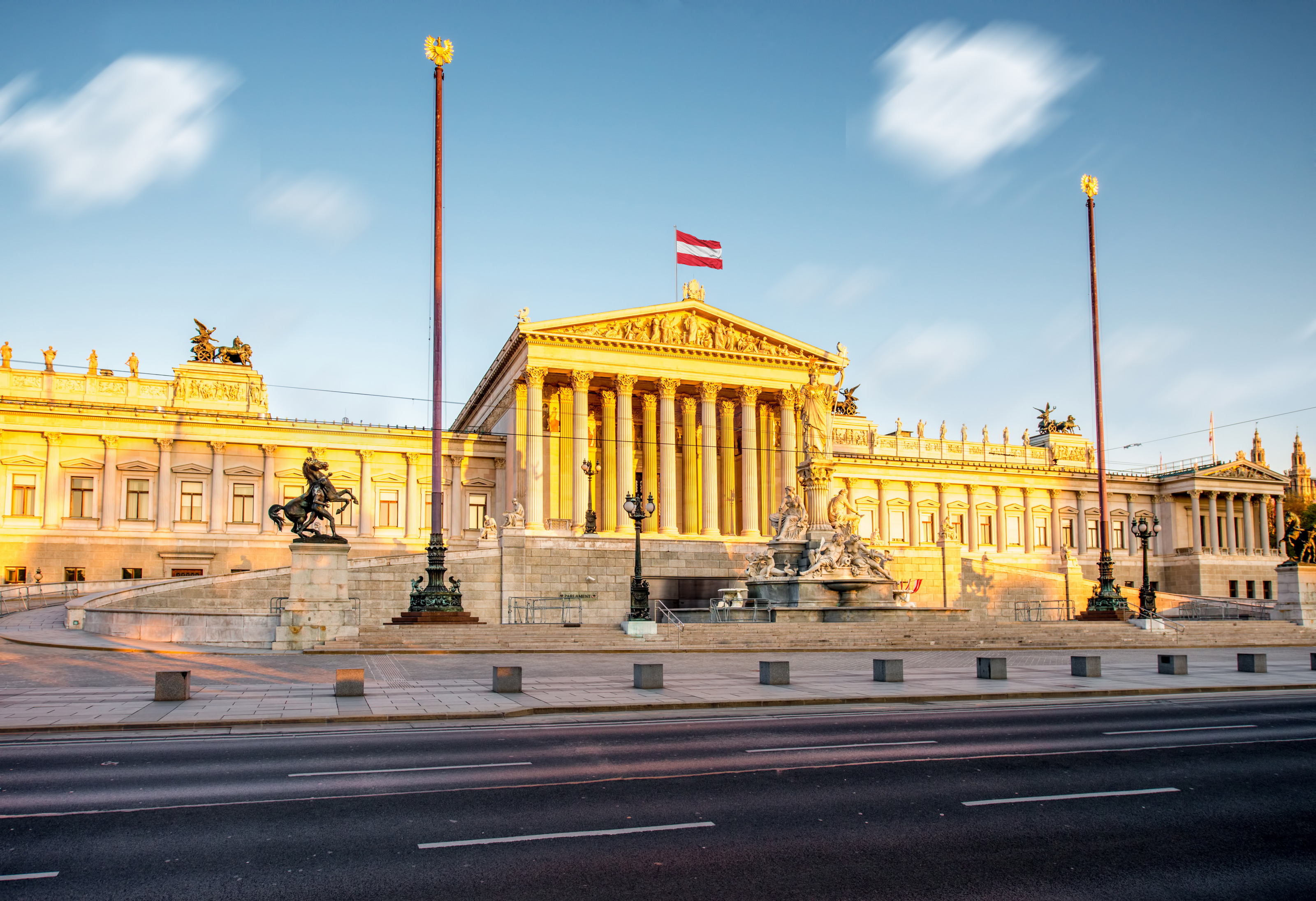 Frontale Aufnahme des Parlaments in Wien mit einer &ouml;sterreichischen Flagge am Dach sowie einem Brunnen vor dem Geb&auml;ude. Der Himmel ist leicht bew&ouml;lkt. Die Sonne scheint auf Teile des Geb&auml;udes. Nirgends sind Menschen