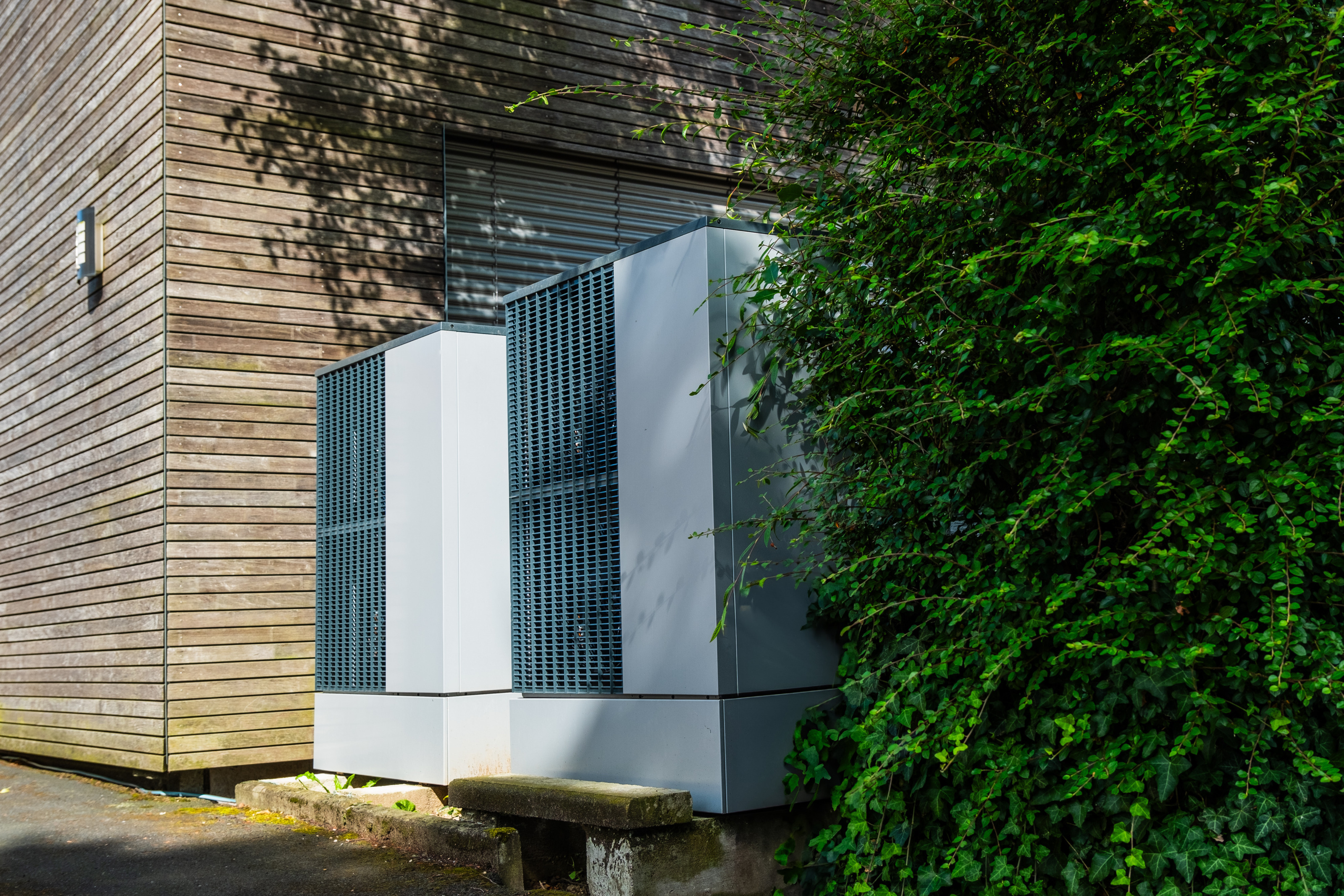 Zwei graue Schr&auml;nke mit Gittern zwischen h&ouml;lzerner Hausfassade und gr&uuml;n belaubter Hecke