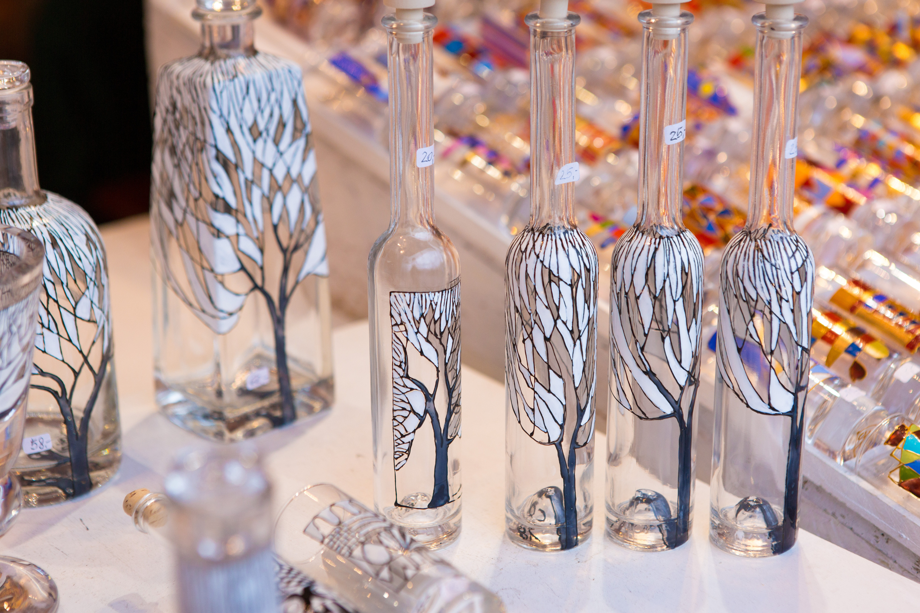 Nahaufnahme mehrerer leerer Flaschen in unterschiedlichen Formen mit hellen Stöpseln und kleinen Zetteln, auf denen Zahlen sind. Jede Flasche ist mit einem Baum mit weißen Blättern bemalt. Im Hintergrund liegen bunt bemalte Gläser
