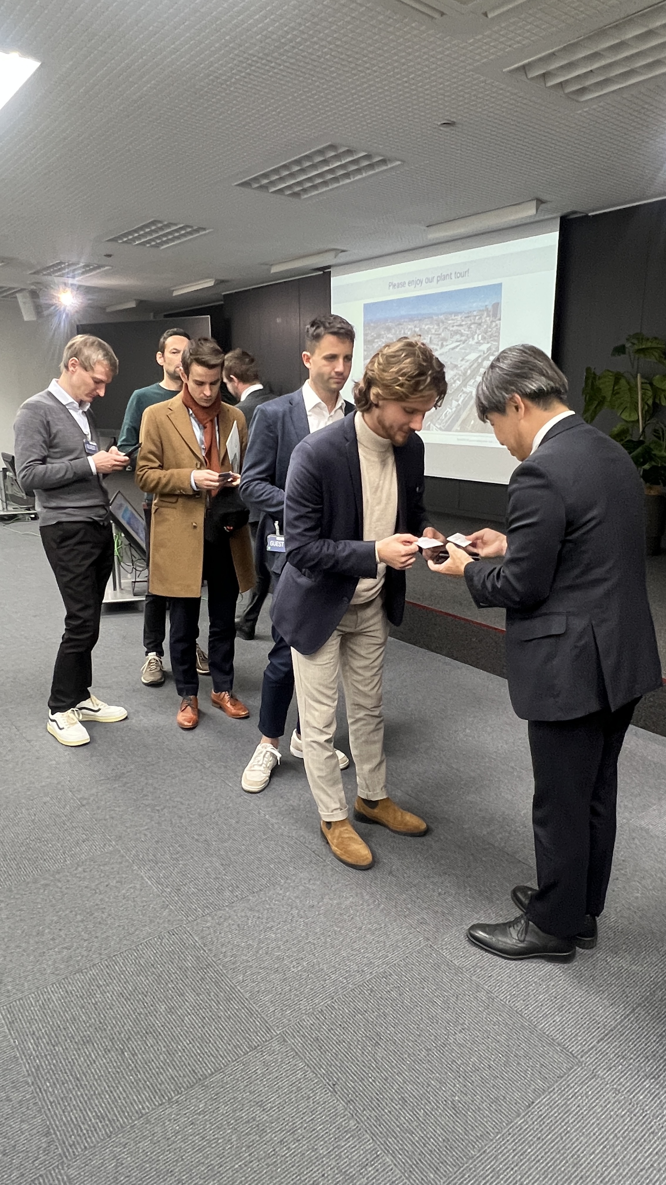 Mehrere Personen bilden eine Schlange in einem Raum. Der ersten Person in der Schlange gegen&uuml;ber steht eine Person in einem Anzug. Die zwei halten jeweils einen Zettel