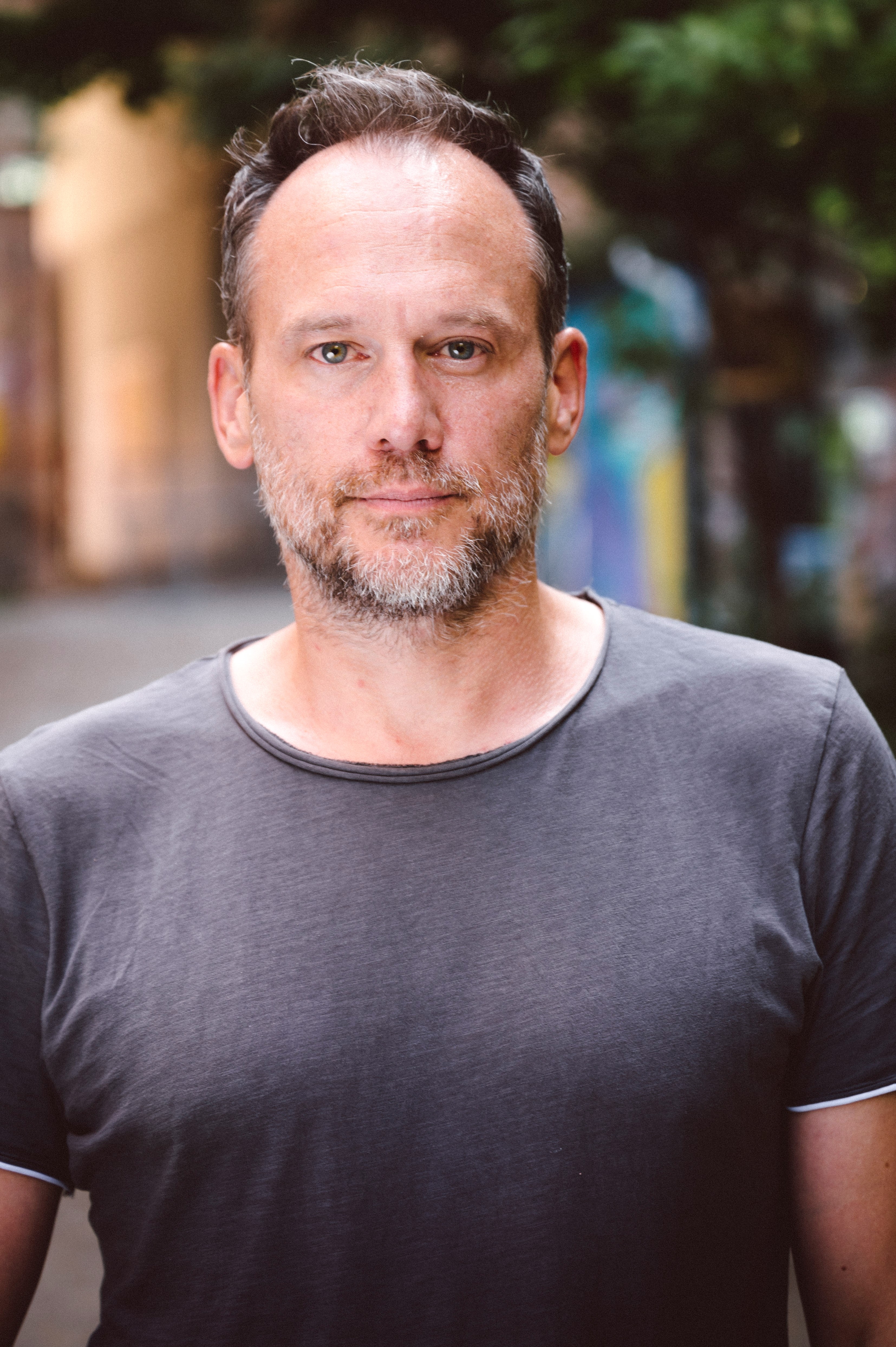 Portr&auml;tfoto einer Person mit kurzem Haar und Bart in grauem T-Shirt vor unscharfem Hintergrund