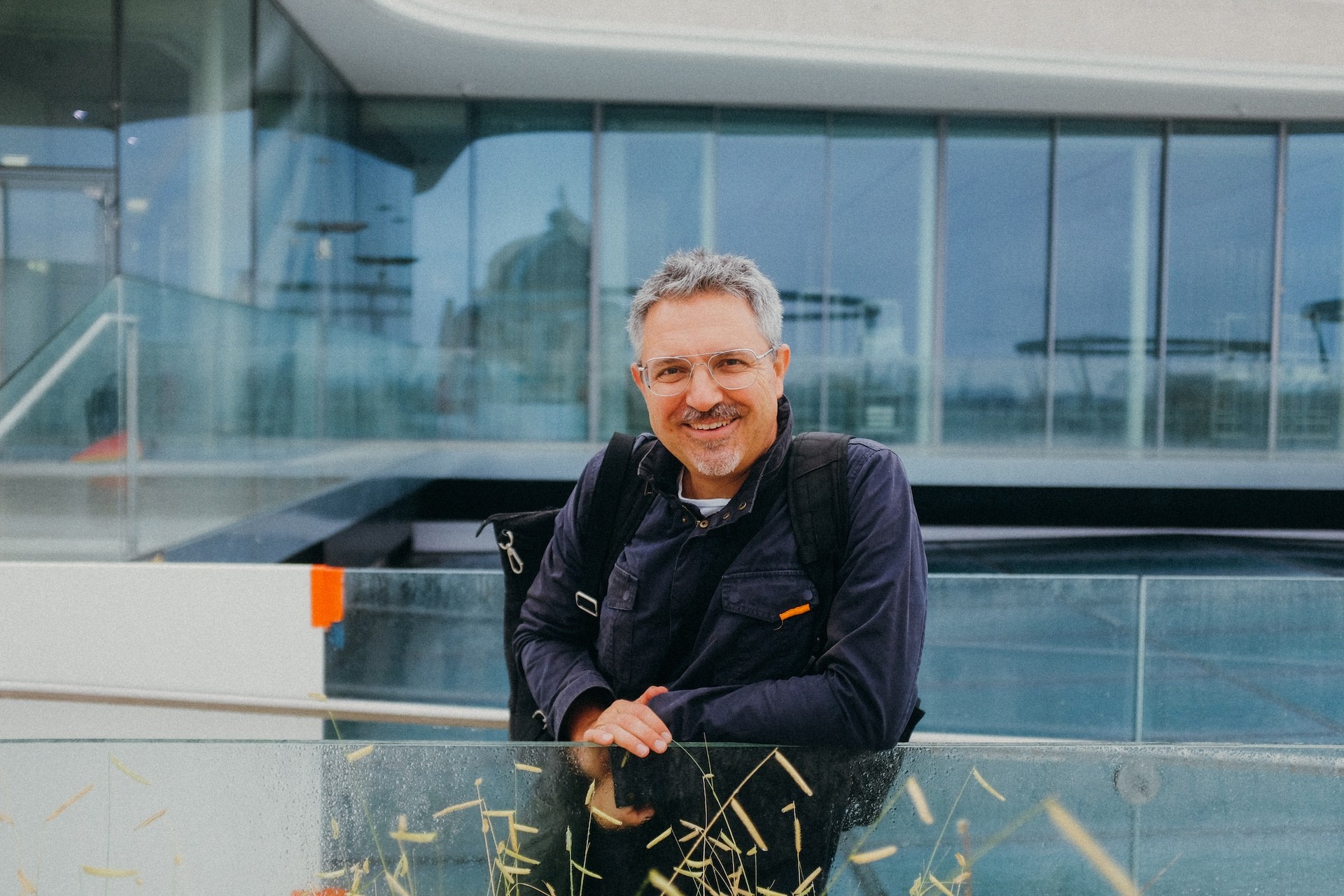 Person mit Brille und Rucksack lehnt an Glasgel&auml;nder in modernem Geb&auml;ude