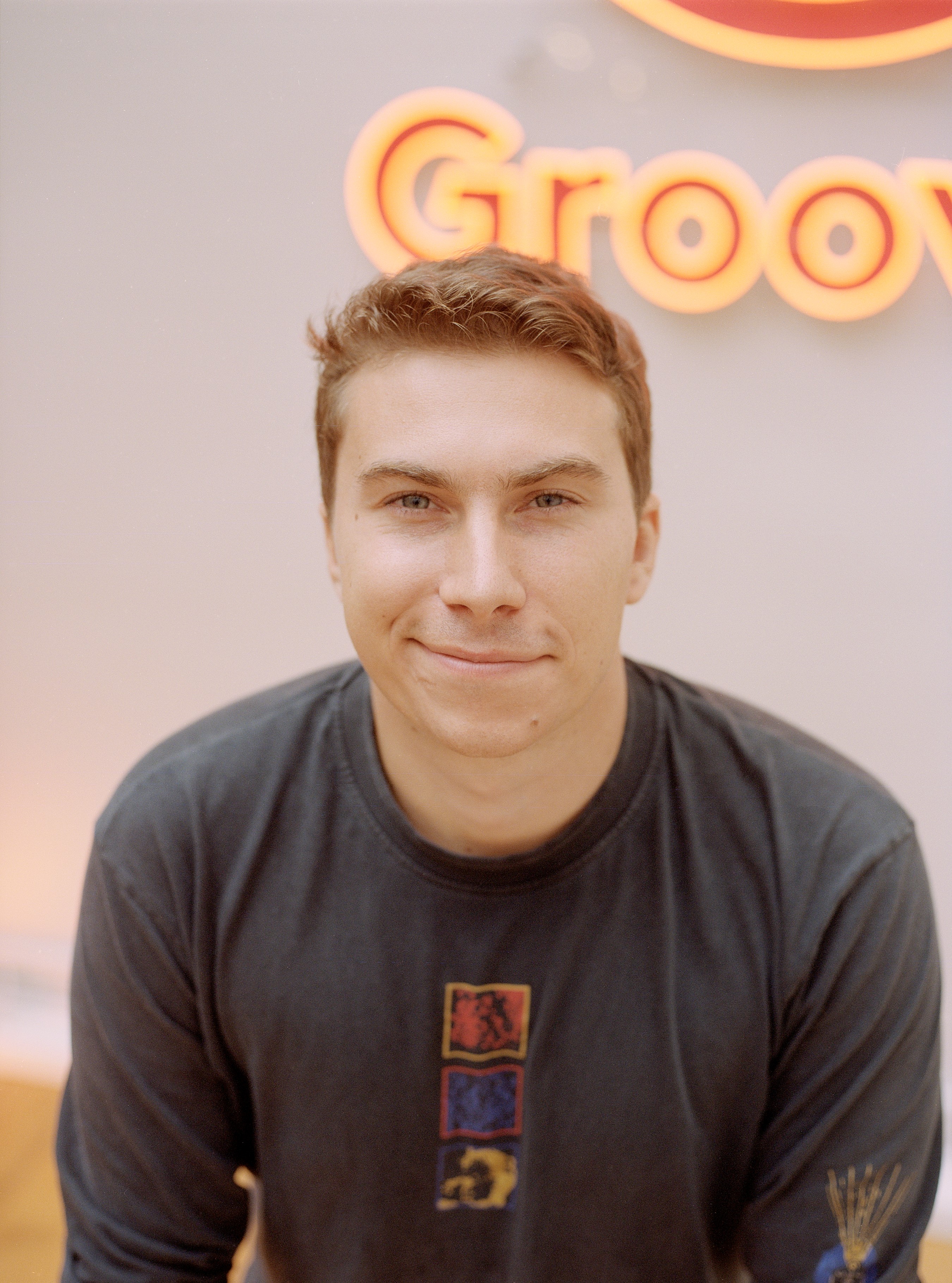 Portr&auml;tfoto: Person mit kurzen hellbraunen Haaren vor Hintergrund mit Schriftzug &bdquo;Groo&ldquo;