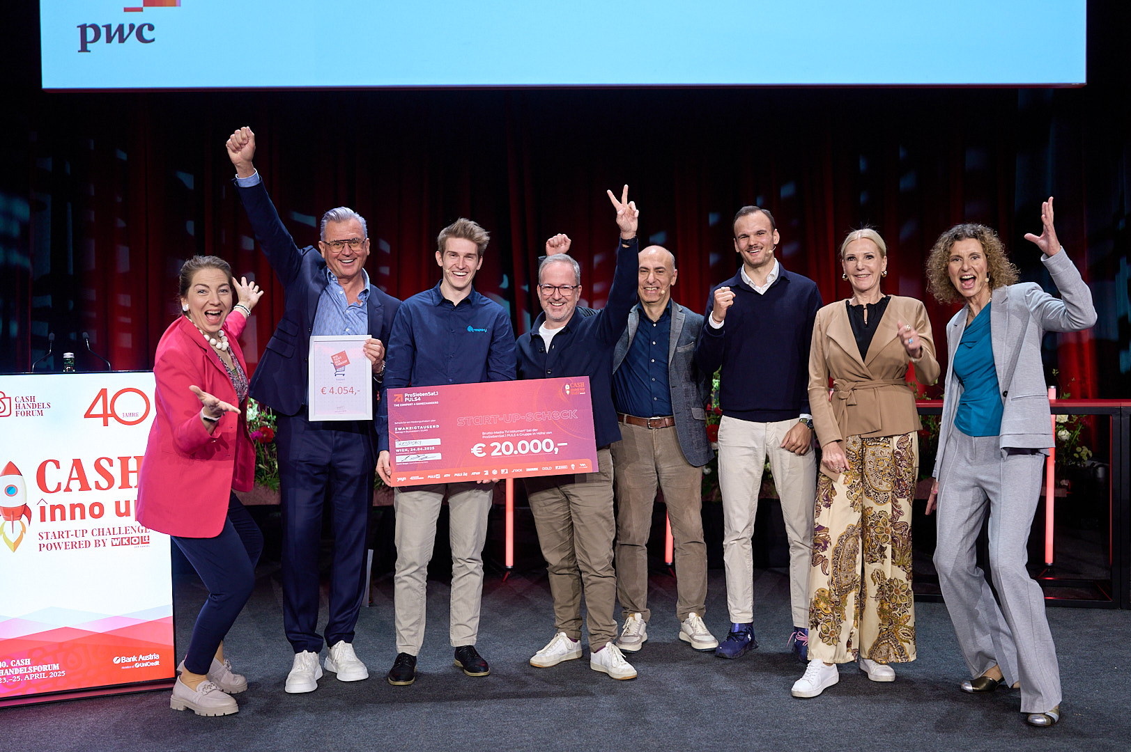 Mehrere Personen stehen nebeneinander auf einer Bühne. In der Mitte ist eine Person, die einen Scheck in Form eines Plakats mit dem Schriftzug € 20.000 in der Hand