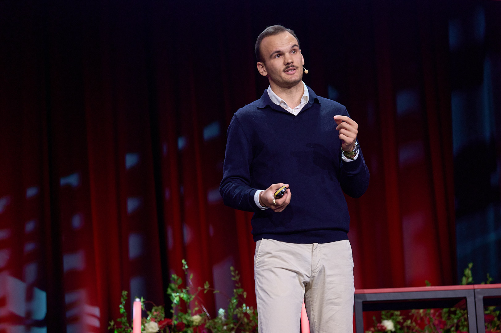 Nahaufnhame einer auf einer Bühne stehenden Person, die in der rechten Hand eine Fernbedienung hat. Mit der linken Hand gestkuliert sie