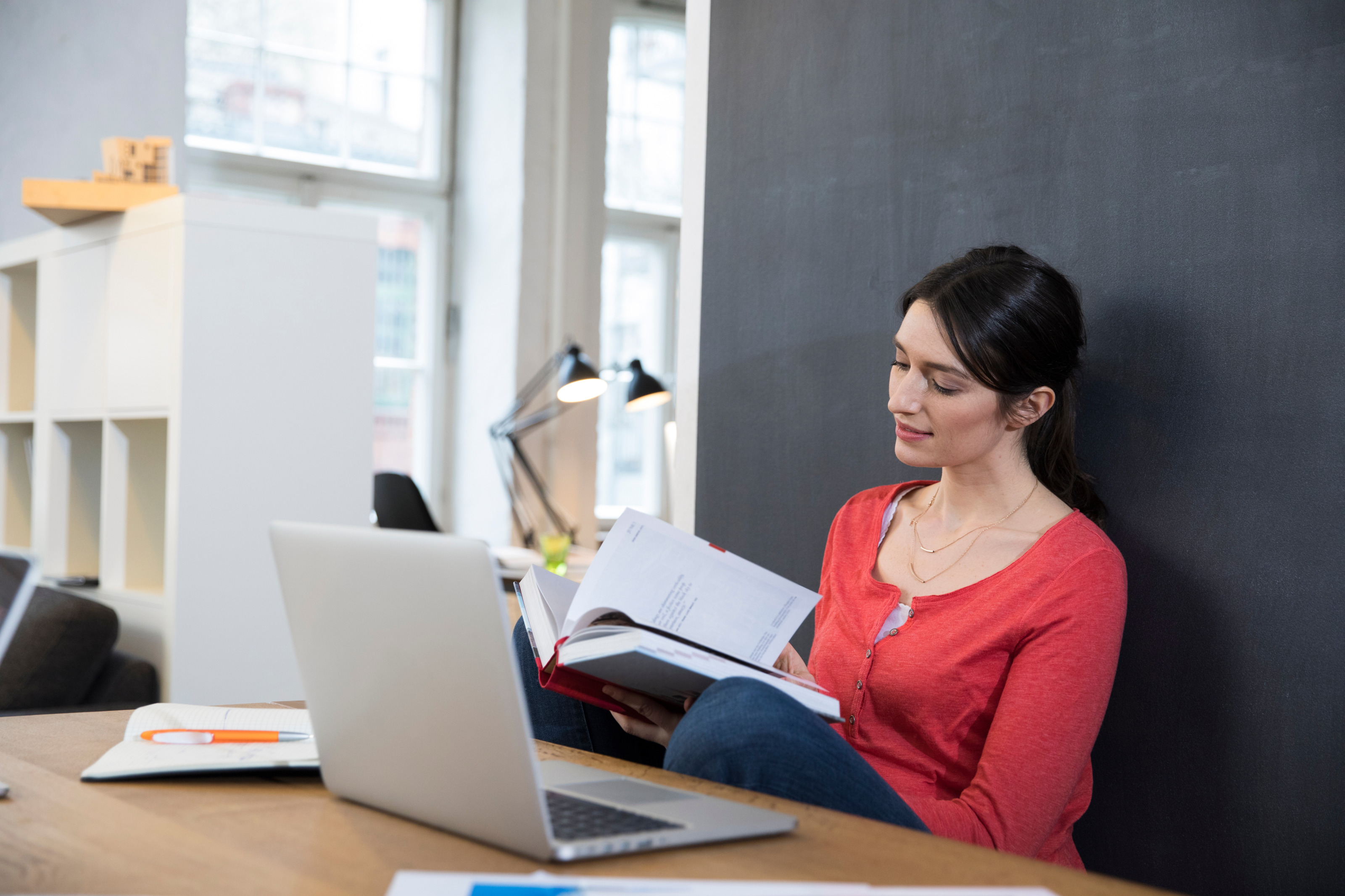 Eine Person sitzt hinter einem Tisch und lehnt sich an einer dunklen Wand an. In den H&auml;nden h&auml;lt sie ein aufgeklapptes Buch, auf das sie blickt. Vor ihr auf dem Tisch steht ein aufgeklappter Laptop