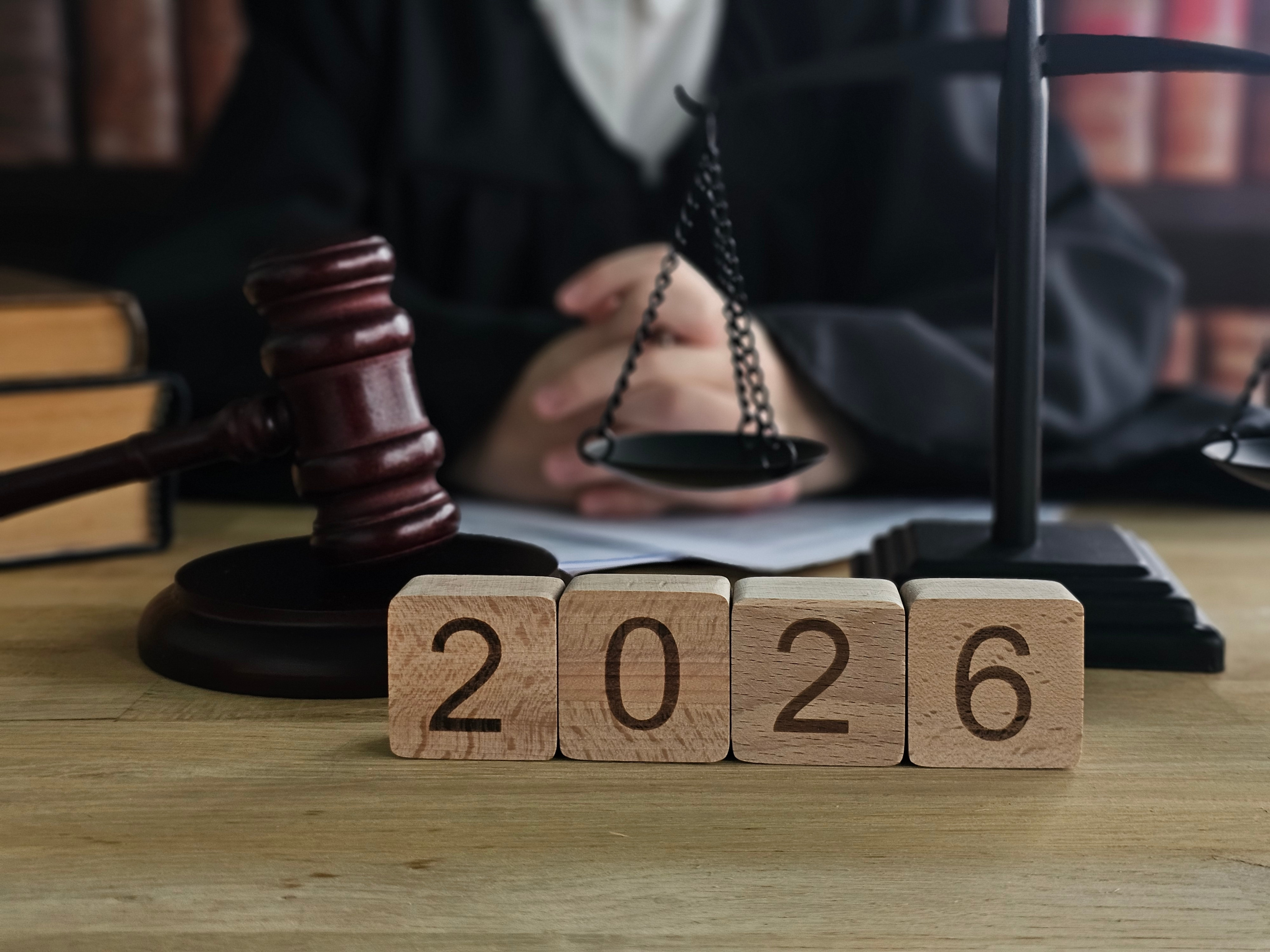 Vier Holzw&uuml;rfel mit dem Schriftzug 2026 im Vordergrund, im Hintergrund ein Richterhammer, eine Waage und eine Person im Ausschnitt an einem Tisch sitzend.