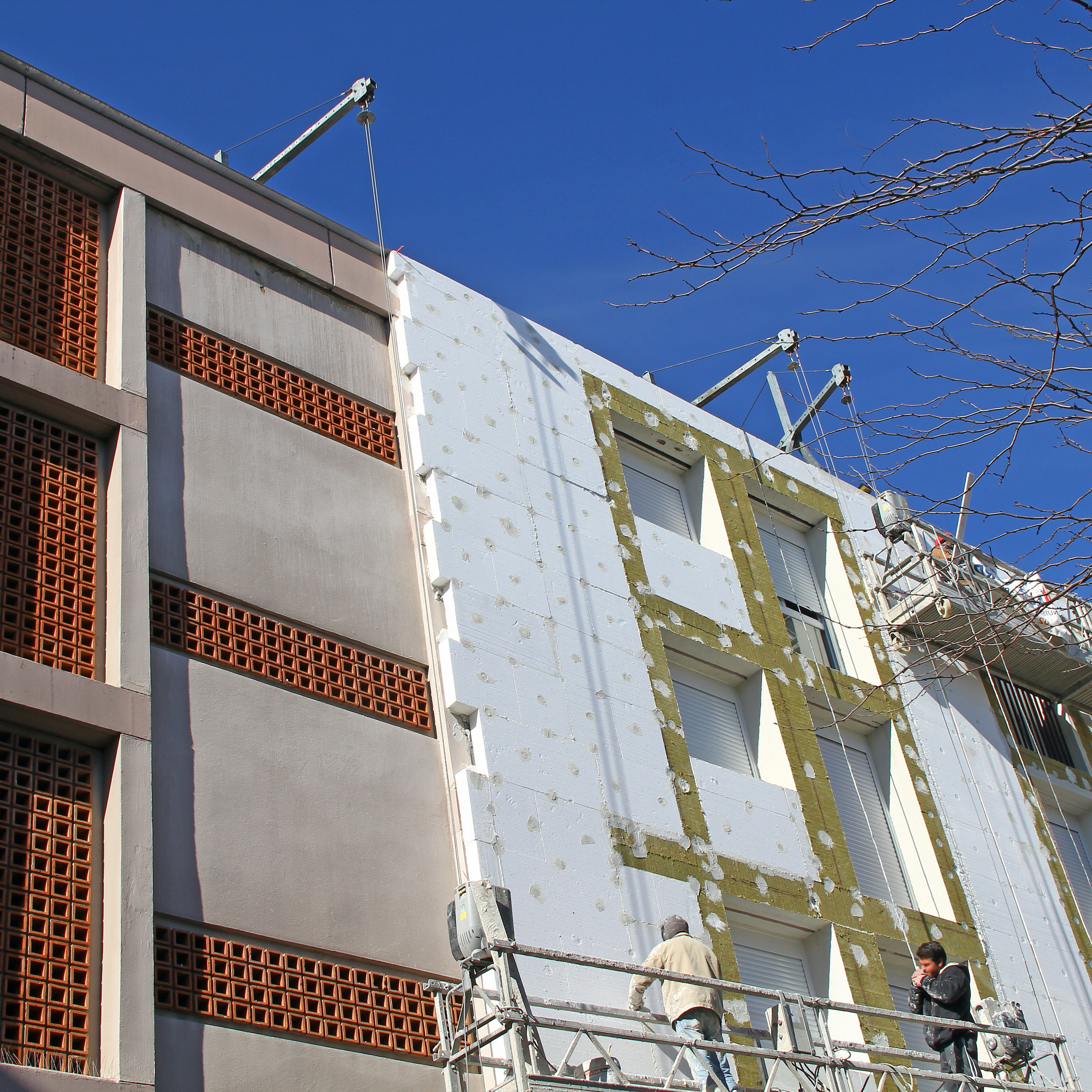 Ein Geb&auml;ude dessen Fassade zur H&auml;lfte mit D&auml;mmmaterialien best&uuml;ckt ist, auf einem Ger&uuml;st stehen zwei Personen.