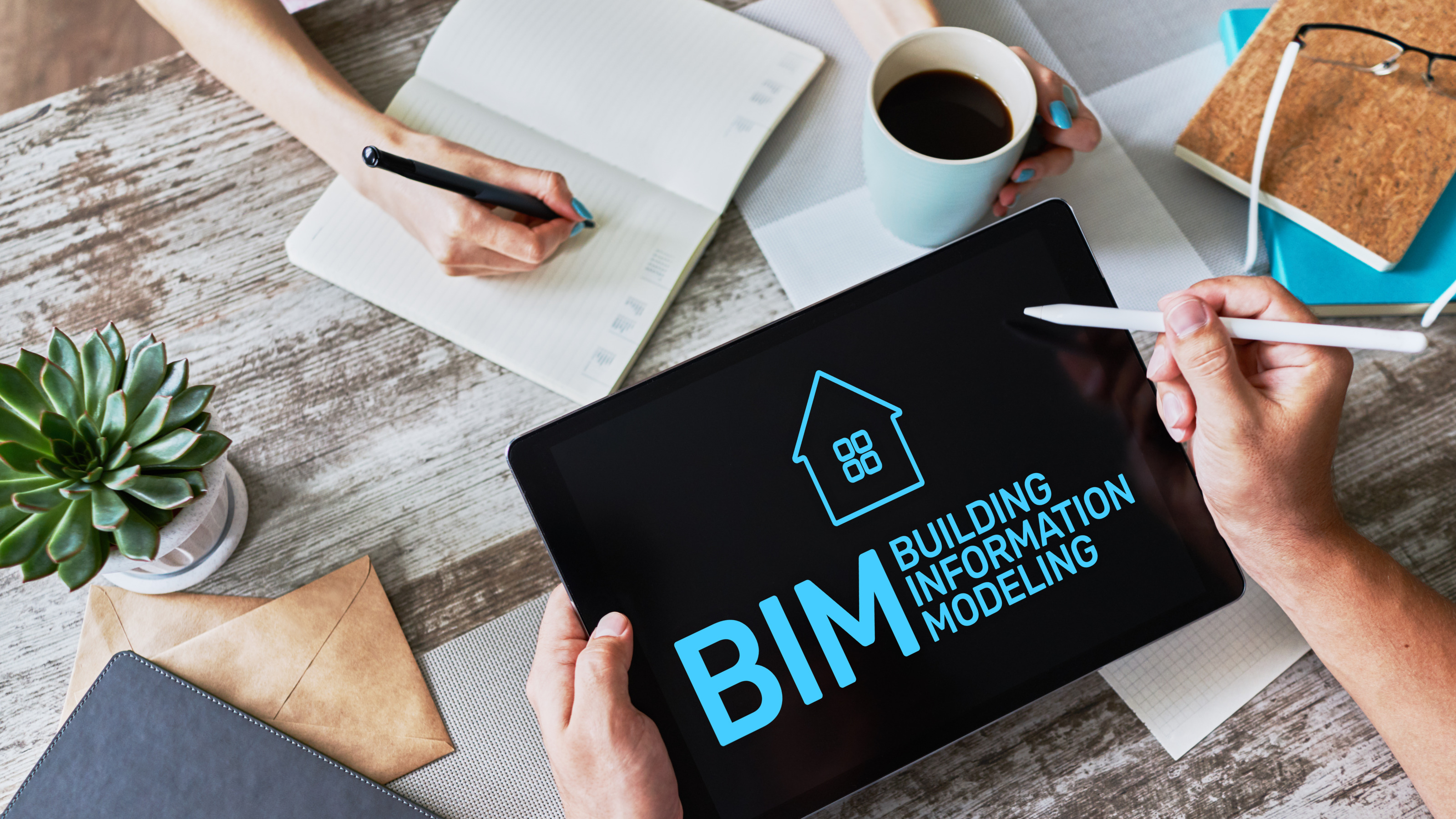 Fokus auf Tablet mit blauer Schrift BIM Building Information Modeling , Tablet wird von H&auml;nden gehalten mit Tabletstift &uuml;ber Tisch mit verschiedenen Schreibtischutensilien,