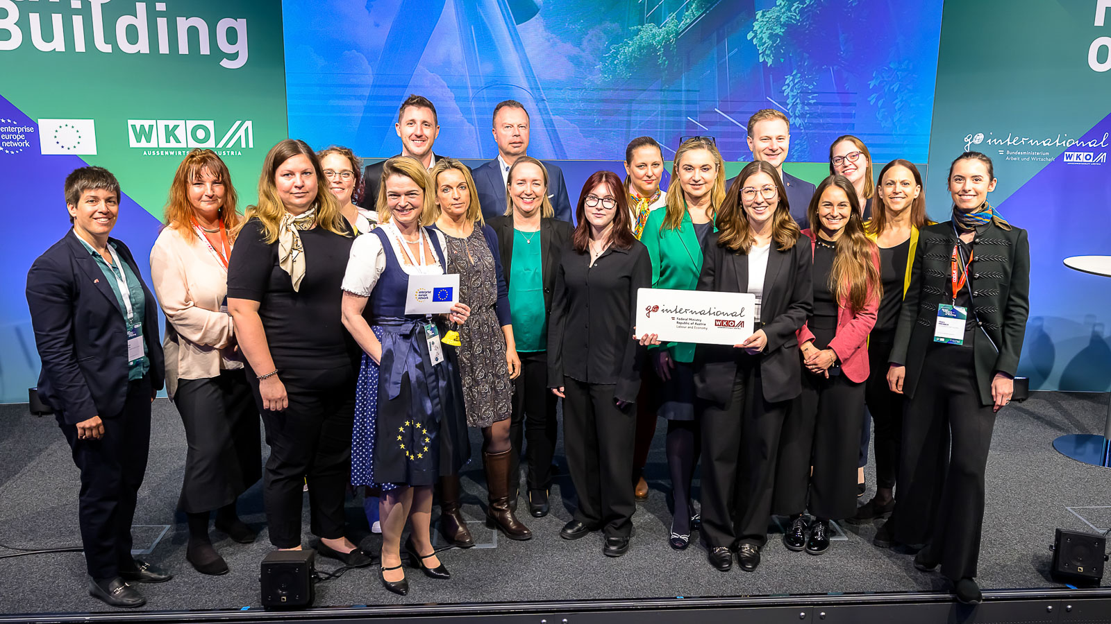 Gruppenbild mehrere Personen neben- und hintereinander vor Wand mit Projektion einer begr&uuml;nten Hausfassade, Logos und Schriftz&uuml;gen wie building, wko und go international stehend