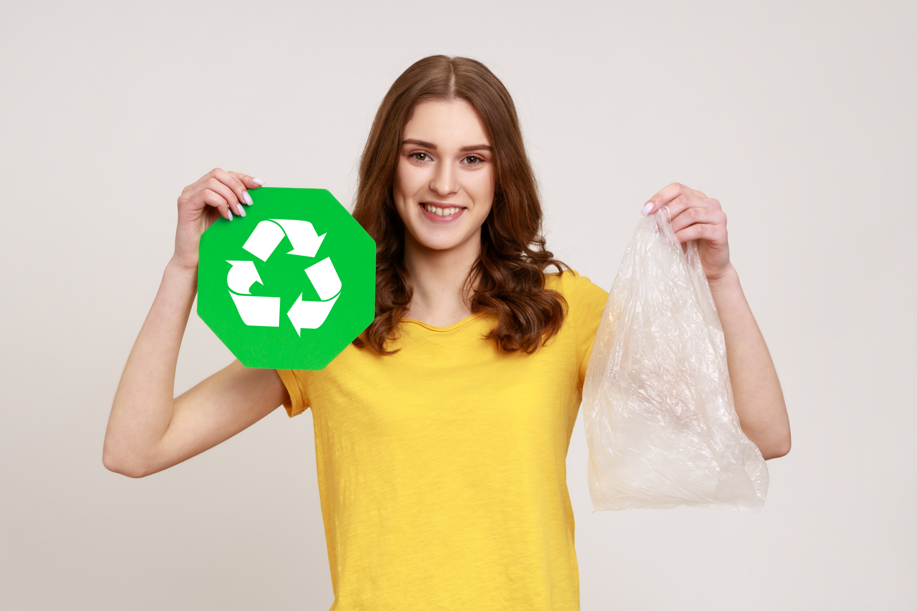 Portrait einer l&auml;chelnden Person in gelbem T-Shirt, die in einer Hand Recyclingzeichen - drei wei&szlig;e im Kreis verlaufenden Pfeile auf wei&szlig;em Hintergrund - und in anderer Hand Plastiksack h&auml;lt