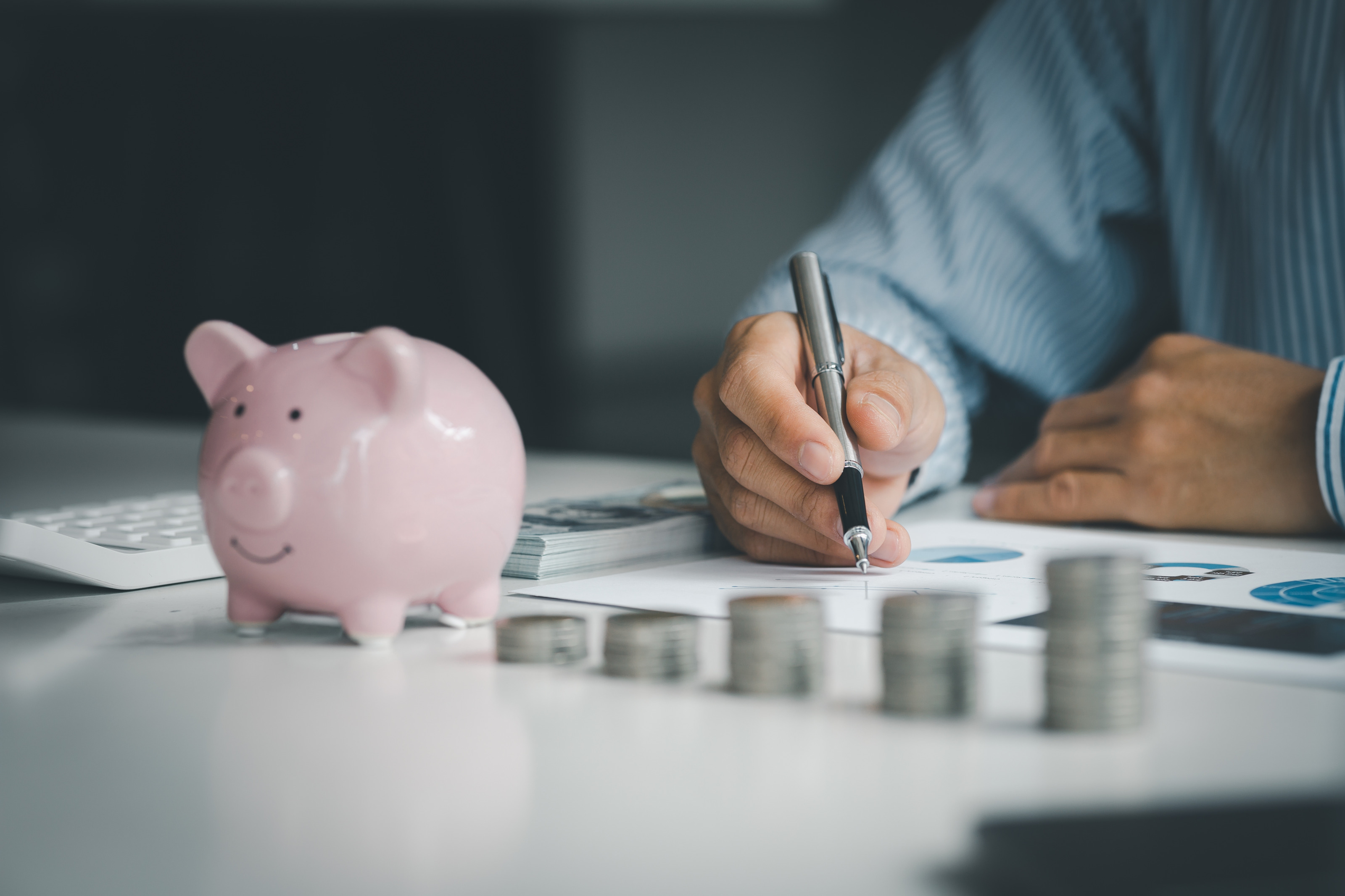 Auf einer Oberfläche sind fünf, unterschiedlich hohe Münzstapel. Links daneben steht eine kleine Figur eines Schweins. Links neben dem Schwein ist ein Taschenrechner. Dahinter sind die Arme einer Person. Unter der rechten Hand sind bedruckte Zettel