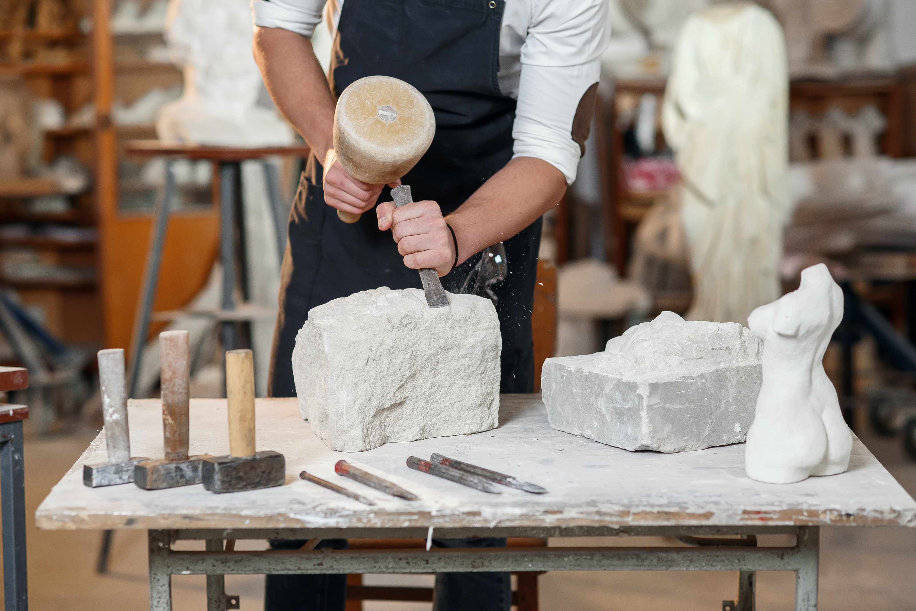 Eine Person mit Sch&uuml;rze im Ausschnitt, die mit Hammer und Mei&szlig;el einen Stein bearbeitet. Auf dem Arbeitstisch befinden sich Steine, eine Steinskulptur, Hammer und Mei&szlig;el. Dahinter erkennt man eine Werkstatt.