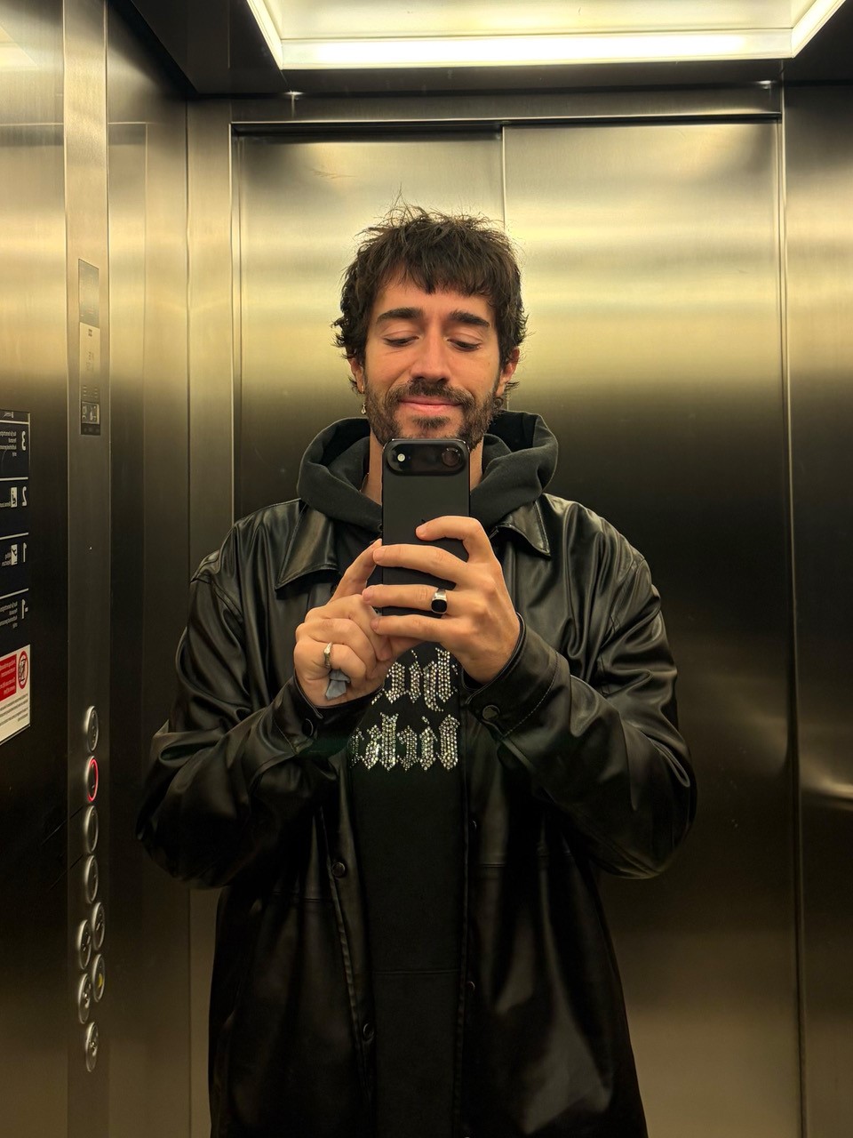 Foto: Person macht Selfie im Aufzug, tr&auml;gt schwarze Jacke und Hoodie, Smartphone vor dem Gesicht