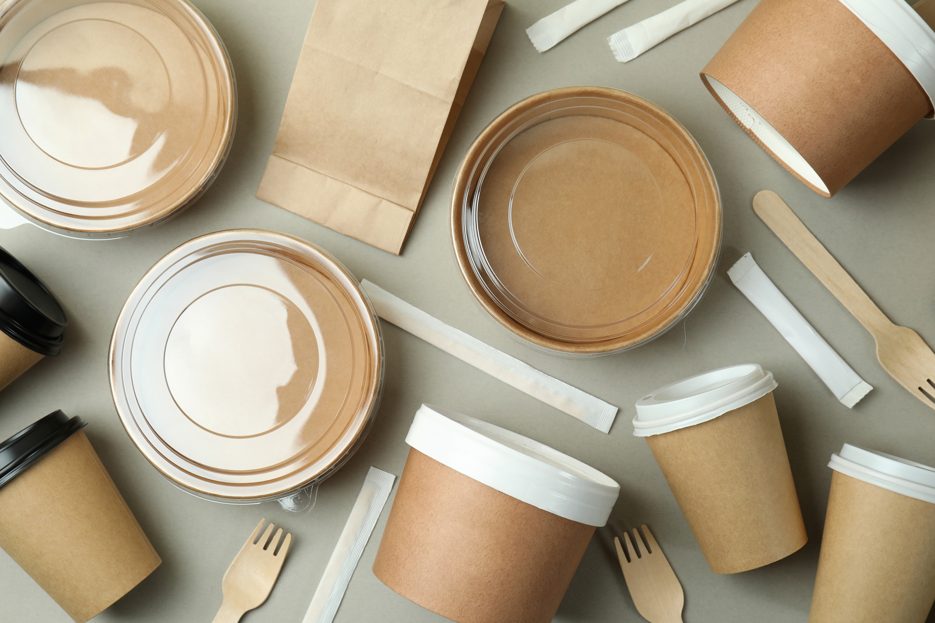 mehrere Becher mit Deckeln, Schalen mit transparenten Deckeln, Papierbeutel, einzeln verpacktes Besteck sowie R&uuml;hrst&auml;bchen. auf hellem Untergrund