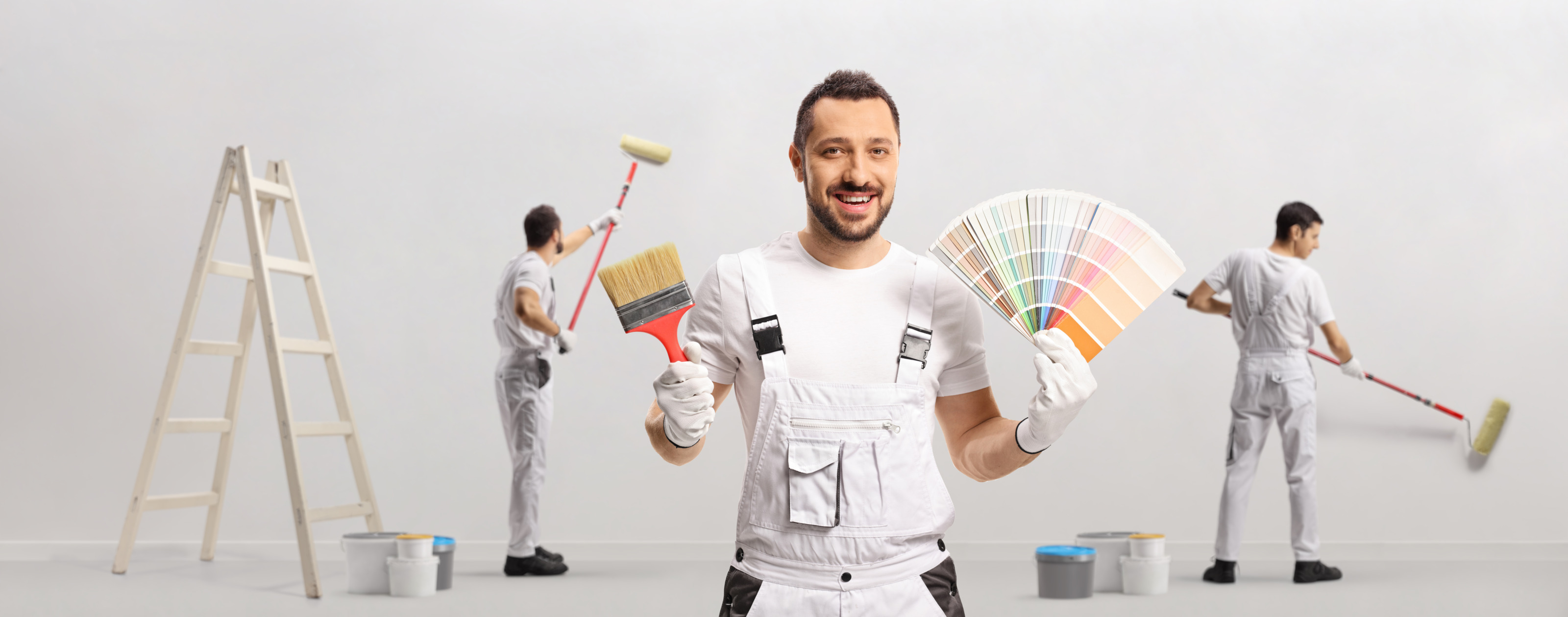Lächelnde Person in weißer Arbeitskleidung hält Malerpinsel sowie Farbfächer in den Händen während zwei weitere Personen im Hintergrund eine Wand ausmalen