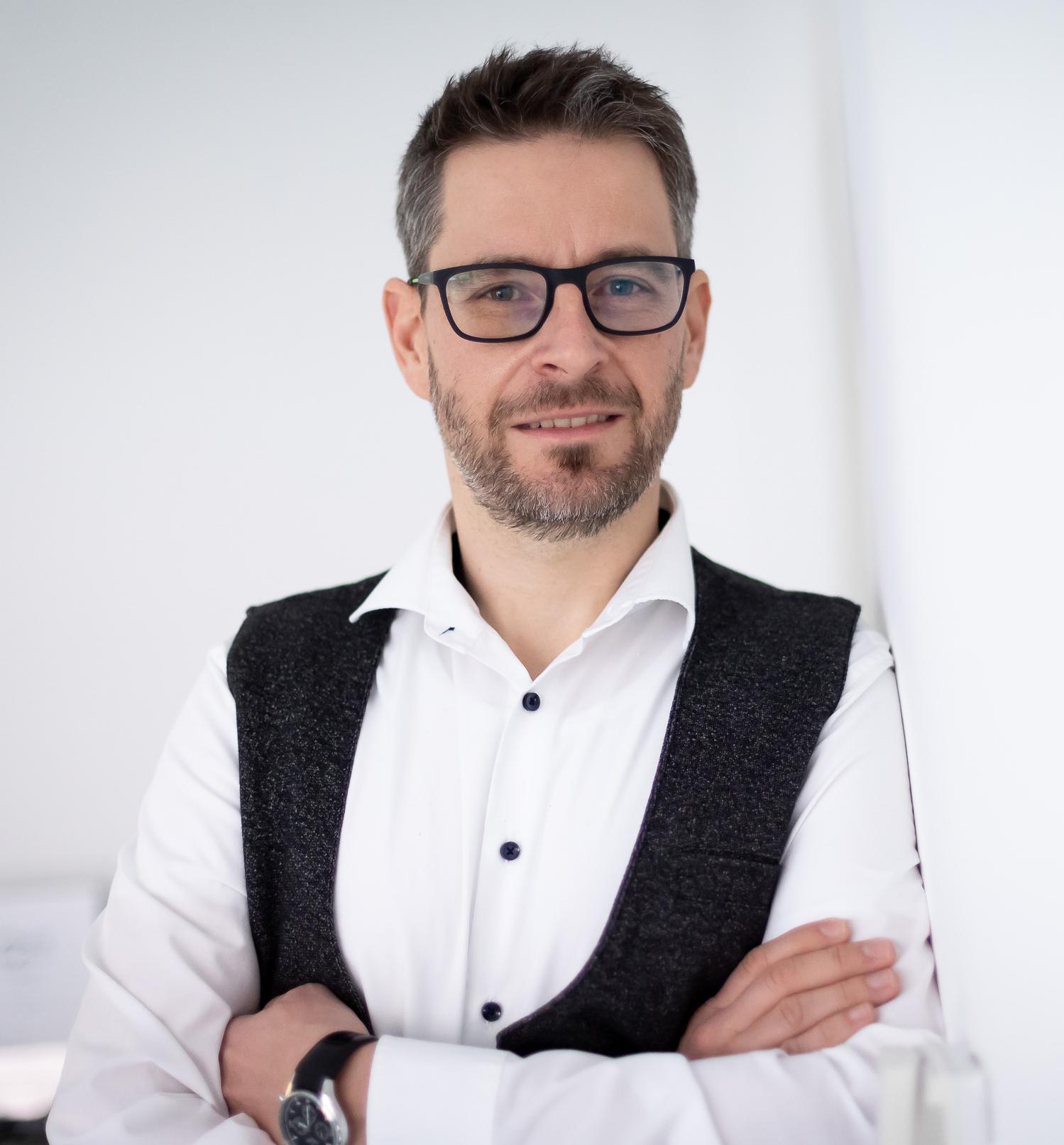 Vortragender Oliver Tatzmann, Mann mit wei&szlig;em Hemd und schwarzem Gillet, hat dunkle kurze Haare, einen Bart und eine Brille, steht vor einem wei&szlig;en Hintergrund