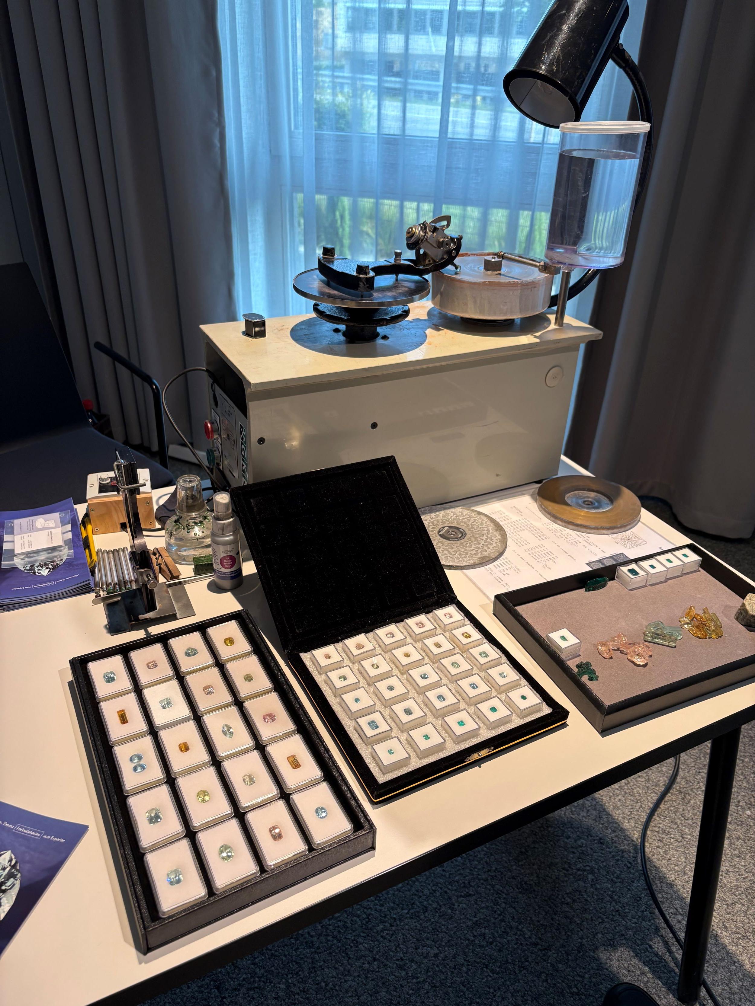 Im Vordergrund zu sehen einzelne Edelsteine in kleinen durchsichtigen Verpackungen, im Hintergrund zu sehen ein Schleifgerät für Edelsteine