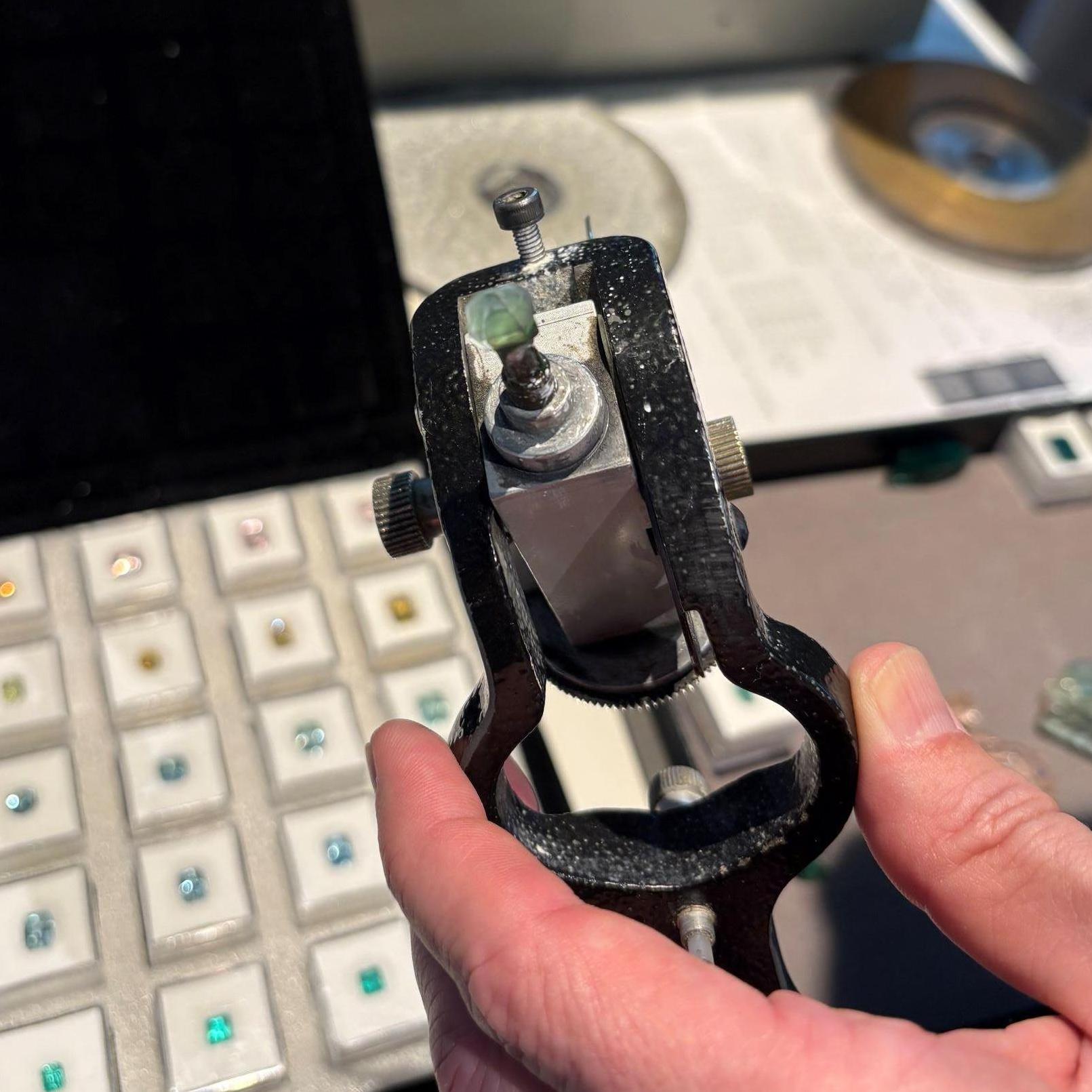 im Vordergrund zu sehen ist ein Edelstein eingespannt in einer Halterung für das Schleifen des Edelsteines, im Hintergrund zu sehen, das Schleifgerät und mehrere Edelsteine