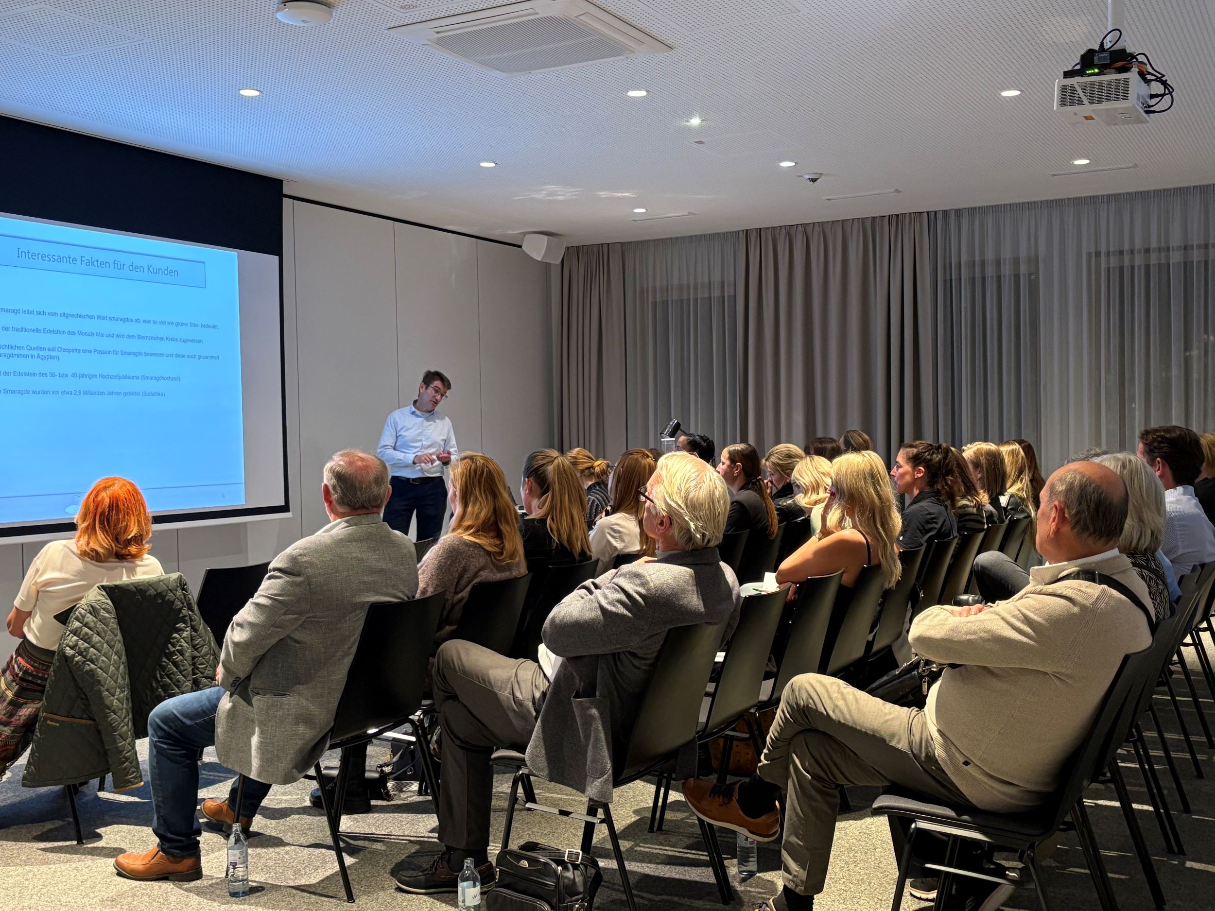 Ein Mann steht vor einer Leinwand, auf der Leinwand ist eine Präsentation zu sehen, im Raum befindet sich noch ein Publikum