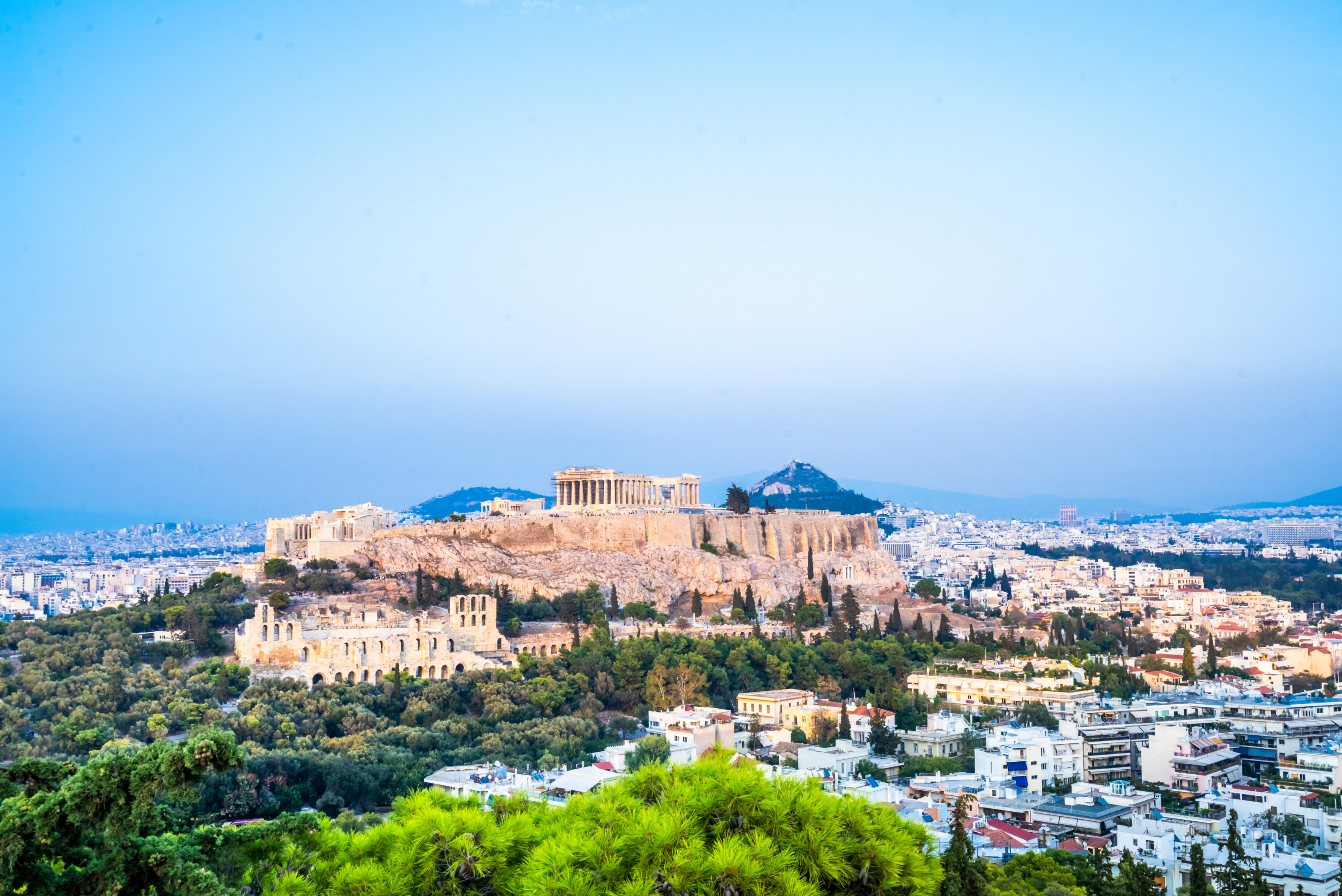 Panoramaaufnahme von Athen mit der Akropolis im Zentrum. Rundherum sind gr&uuml;ne B&auml;ume so wie sehr viele Wohngeb&auml;ude. Im Hintergrund sind H&uuml;gel