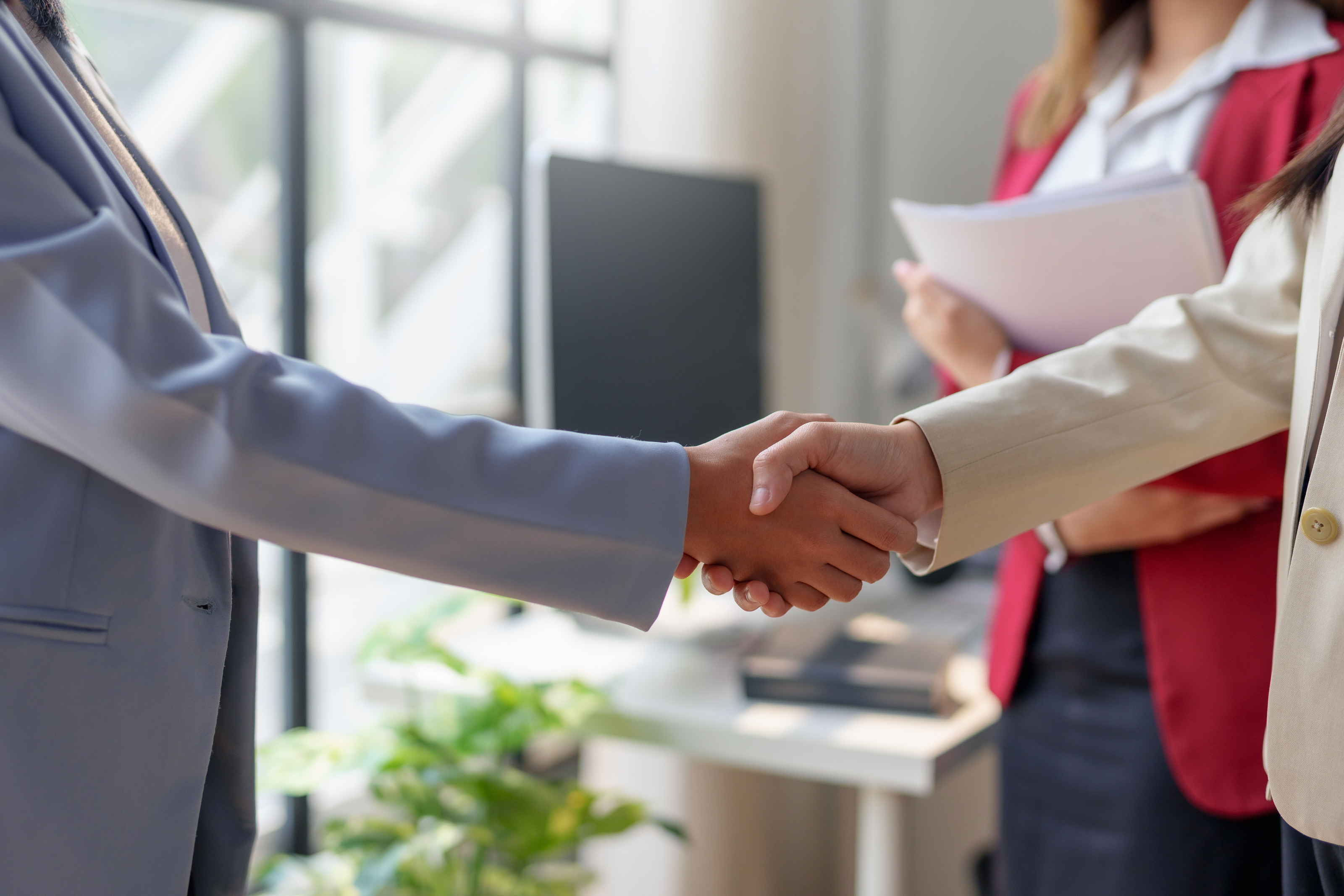 Seitliche Aufnahme zweier Personen in Sakkos, die sich gegen&uuml;ber stehen und die H&auml;nde sch&uuml;tteln. Neben ihnen steht eine weitere Person, die einen Stapel Zettel h&auml;lt