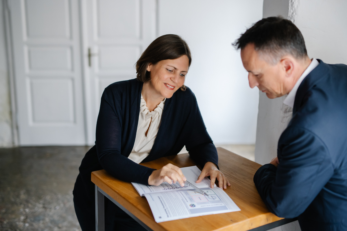 Gudrun Gruber ber&auml;t einen Mann indem sie ihm etwas auf einem Blatt zeigt. Sie sitzen einander gegen&uuml;ber.