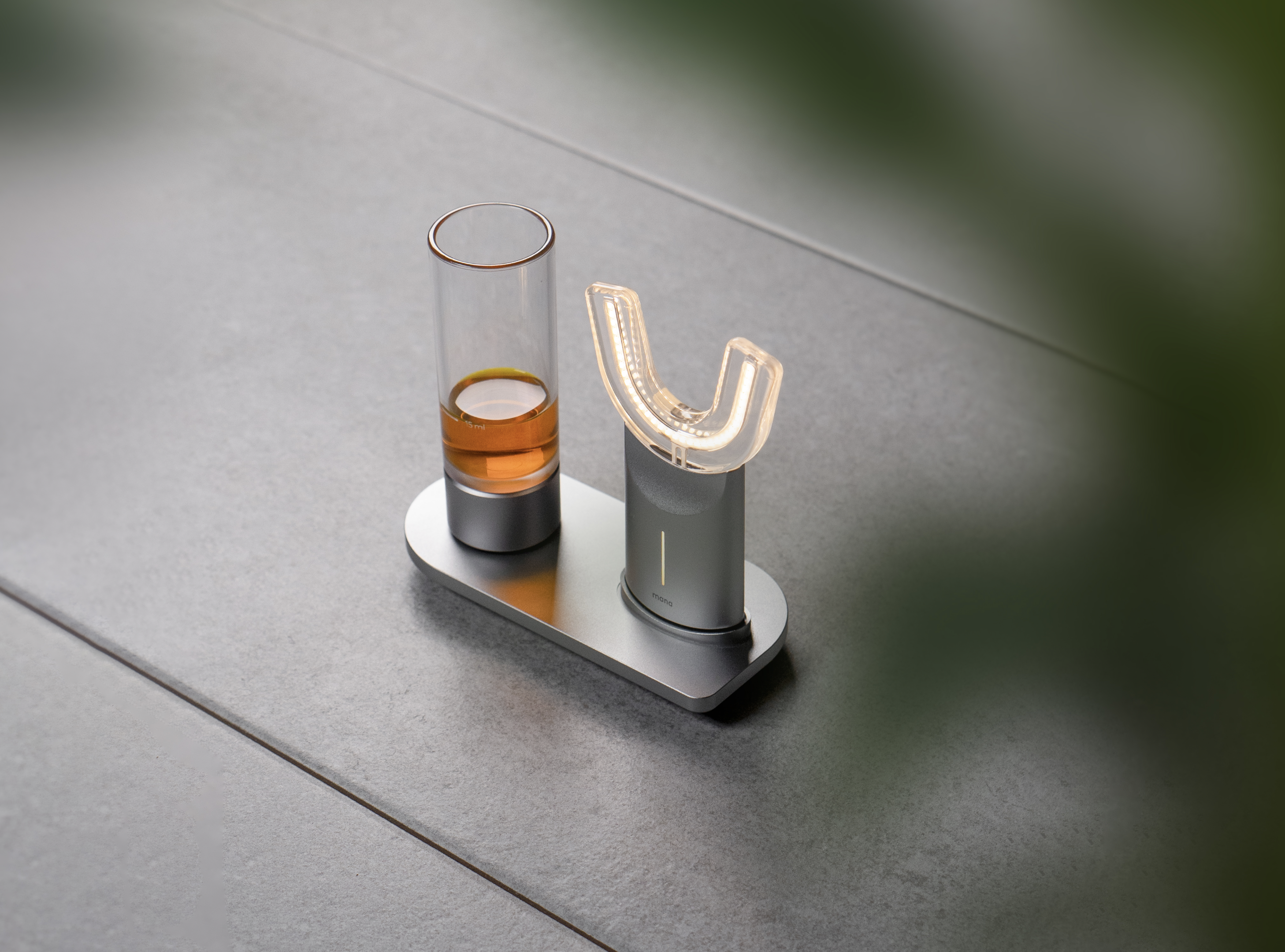 Braune Flüssigkeit in Stamperl, daneben Zahnlicht in Kieferform, alles auf einer grauen Ladestation fix verankert