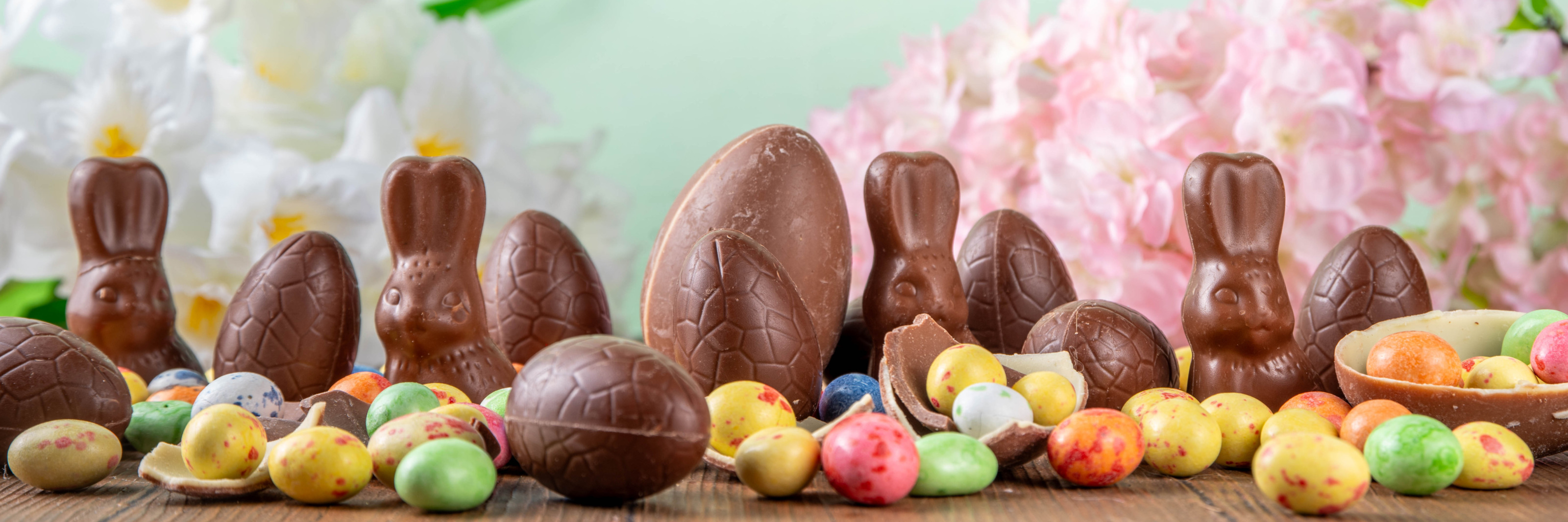 Schokoladeneier, Schokoladenhasen und bunte Eier liegen quer verstreut auf Holzbrettern. Im Hintergrund sind Blumen