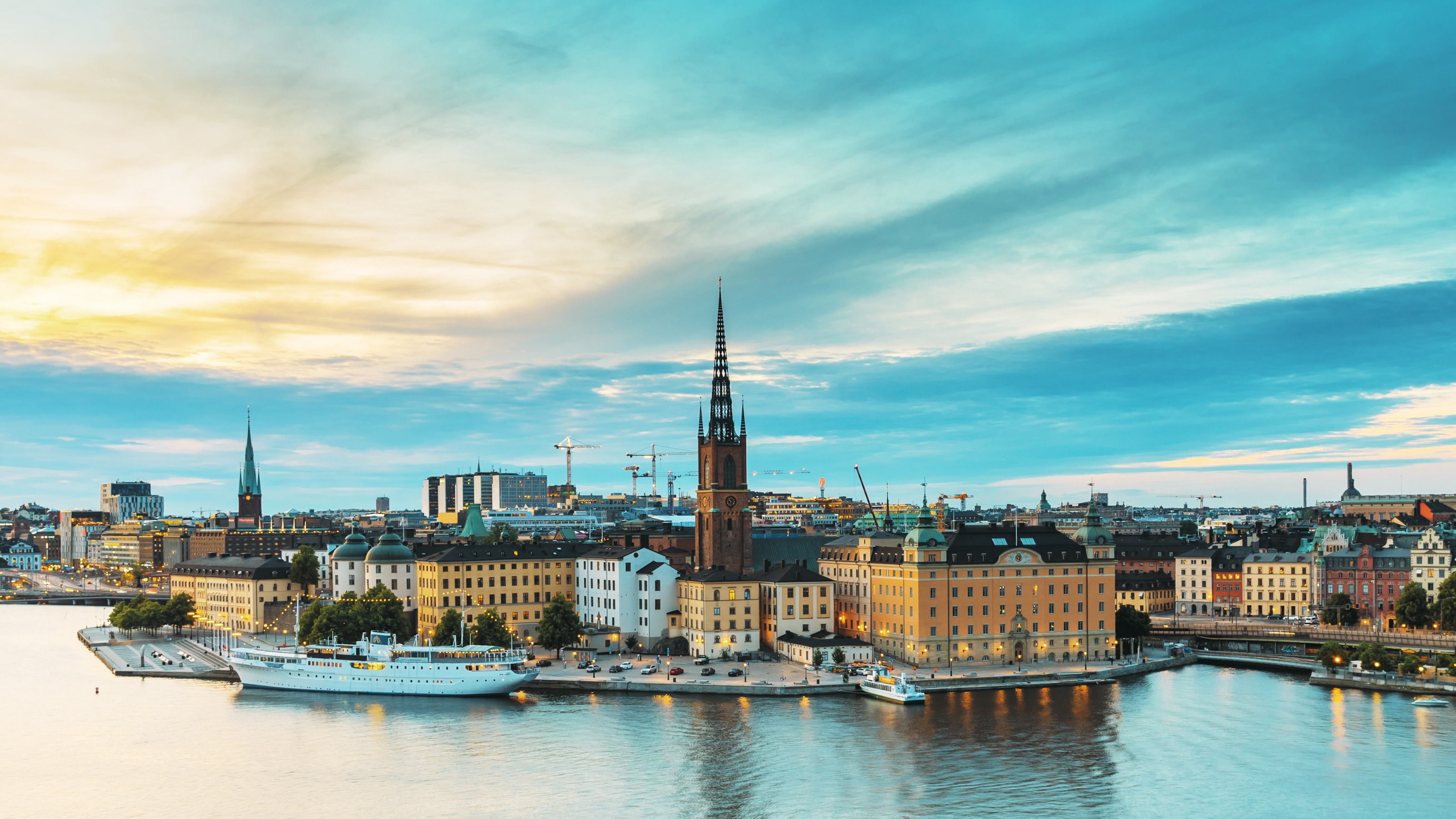 Panoramaaufnahme des historischen Zentrums der Stadt Stockholm mit verschiedenen Geb&auml;uden, Kircht&uuml;rmen, Kr&auml;nen und einem Gew&auml;sser im Vordergrund. Auf dem Gew&auml;sser sind teilweise Boote
