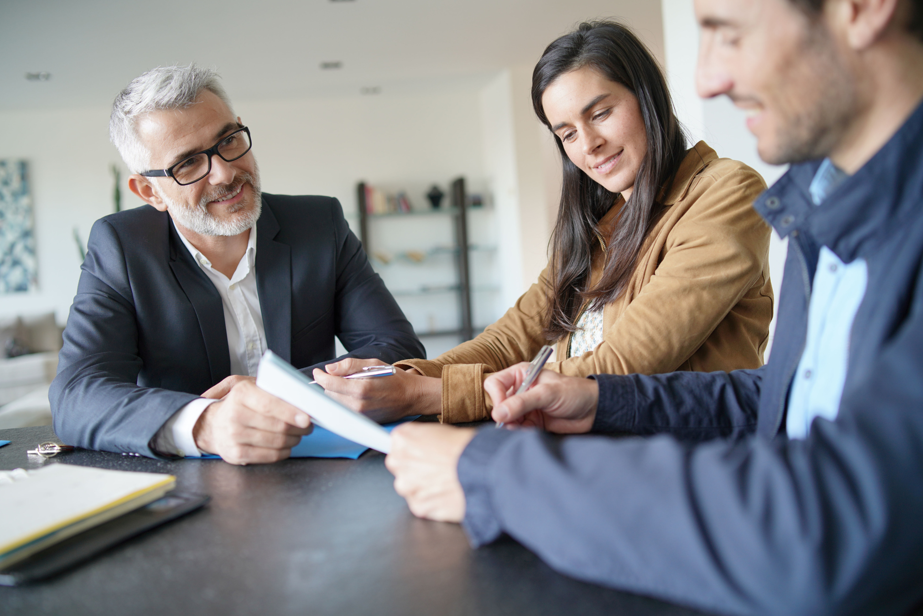 Drei Personen an Tisch sitzend, zwei haben Stifte in der Hand, eine der Personen unterzeichnet Dokument.