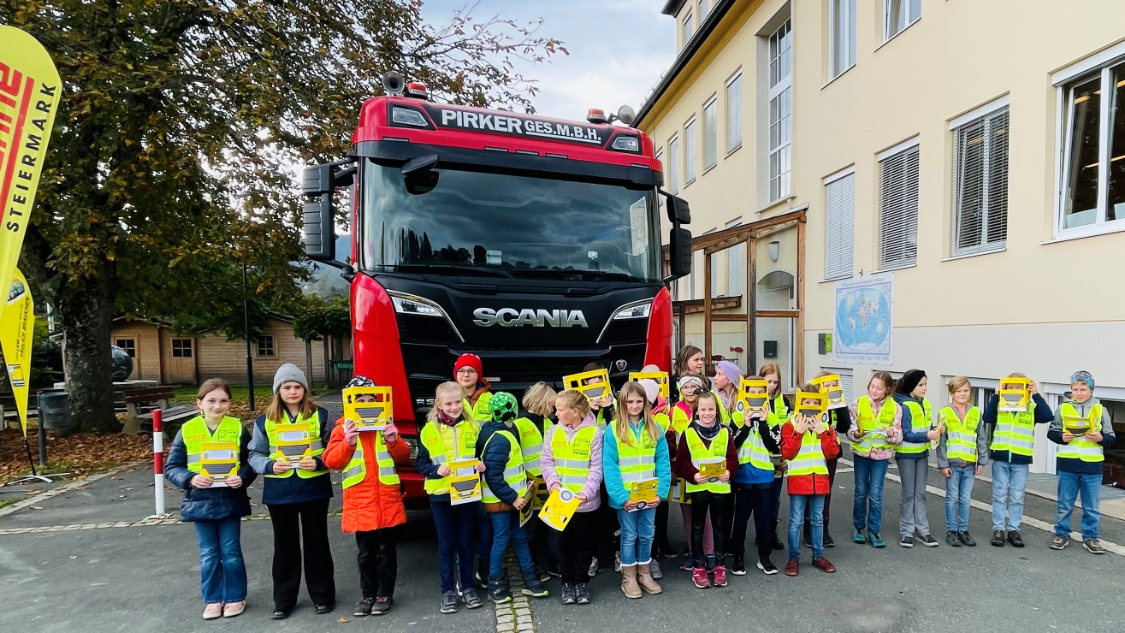 Eine Gruppe Kinder steht in gelben Warnwesten vor einem roten LKW und hält LKW-Modelle aus gelbem Papier hoch