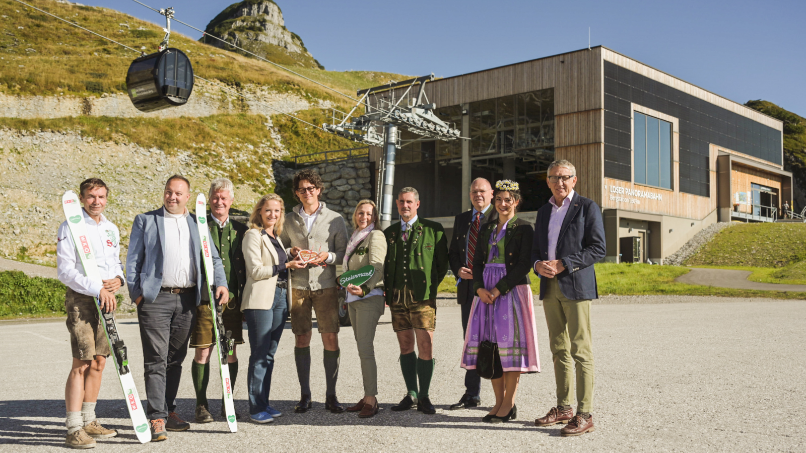 Gruppenfoto von Männern und Frauen - teils in Tracht, teils im Businessoutfit - vor der Talstation einer Seilbahn, links oben sieht man eine Gondel