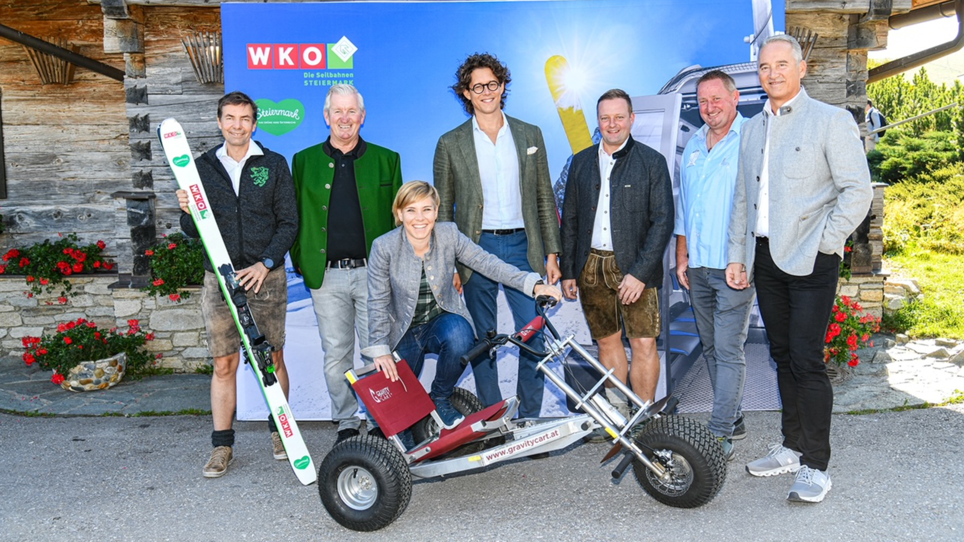 6 Herren und 1 Dame posieren vor einem großen himmelblauen WKO Poster vor einer Hütte am Berg; einer hält Skier in der Hand und die Dame stützt sich auf ein dreirädriges Gefährt (gravitycart)