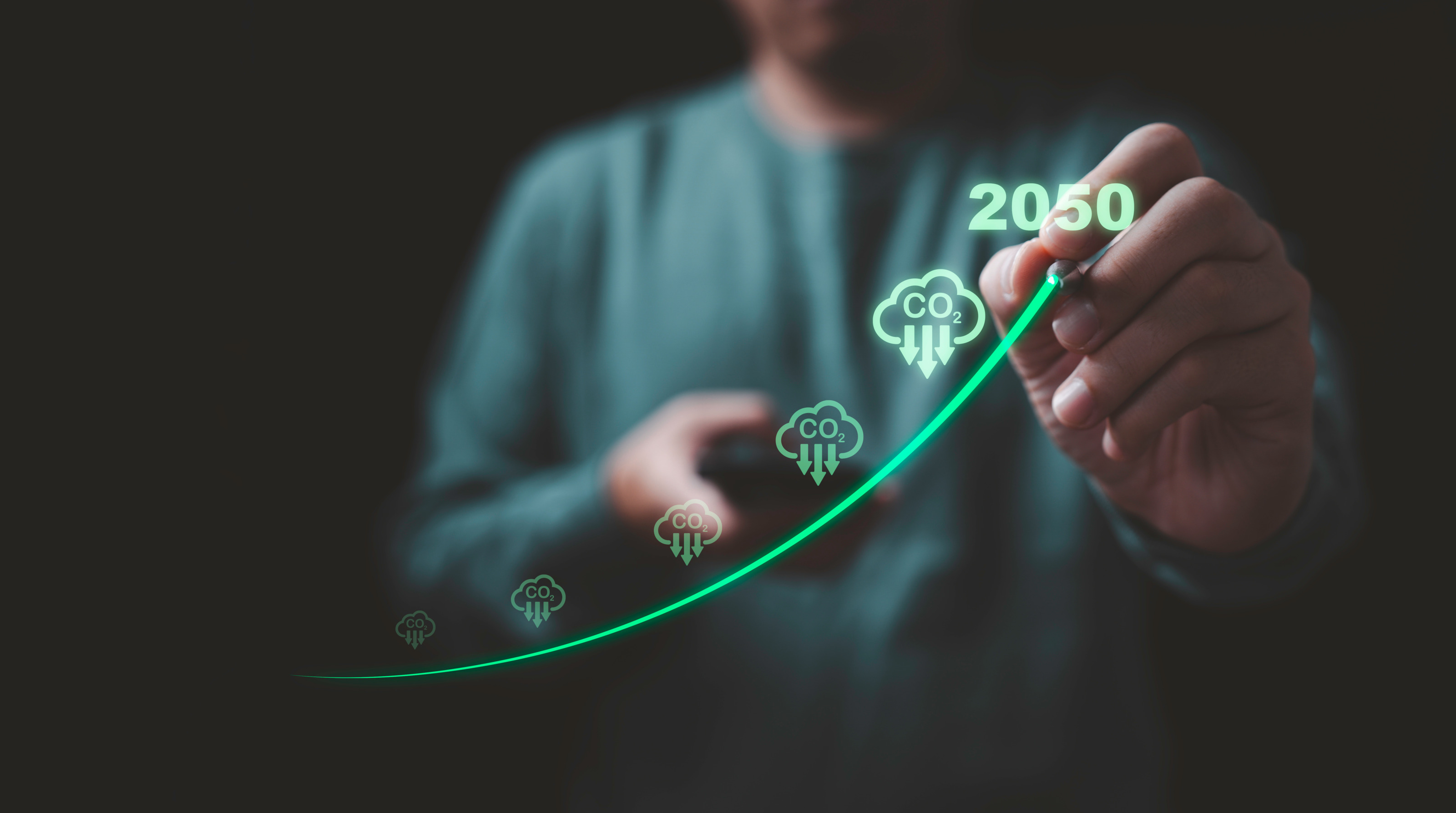 Person in gr&uuml;nem Pullover im Ausschnitt im Hintergrund verschwommen Arm nach vorne reichend, Hand im Fokus gr&uuml;n leuchtende nach oben geschwungenen Linie in Luft zeichnend mit Wolken mit Schrift CO2 und Pfeilen nach unten, zu oberst gr&uuml;n leuchtend 2050