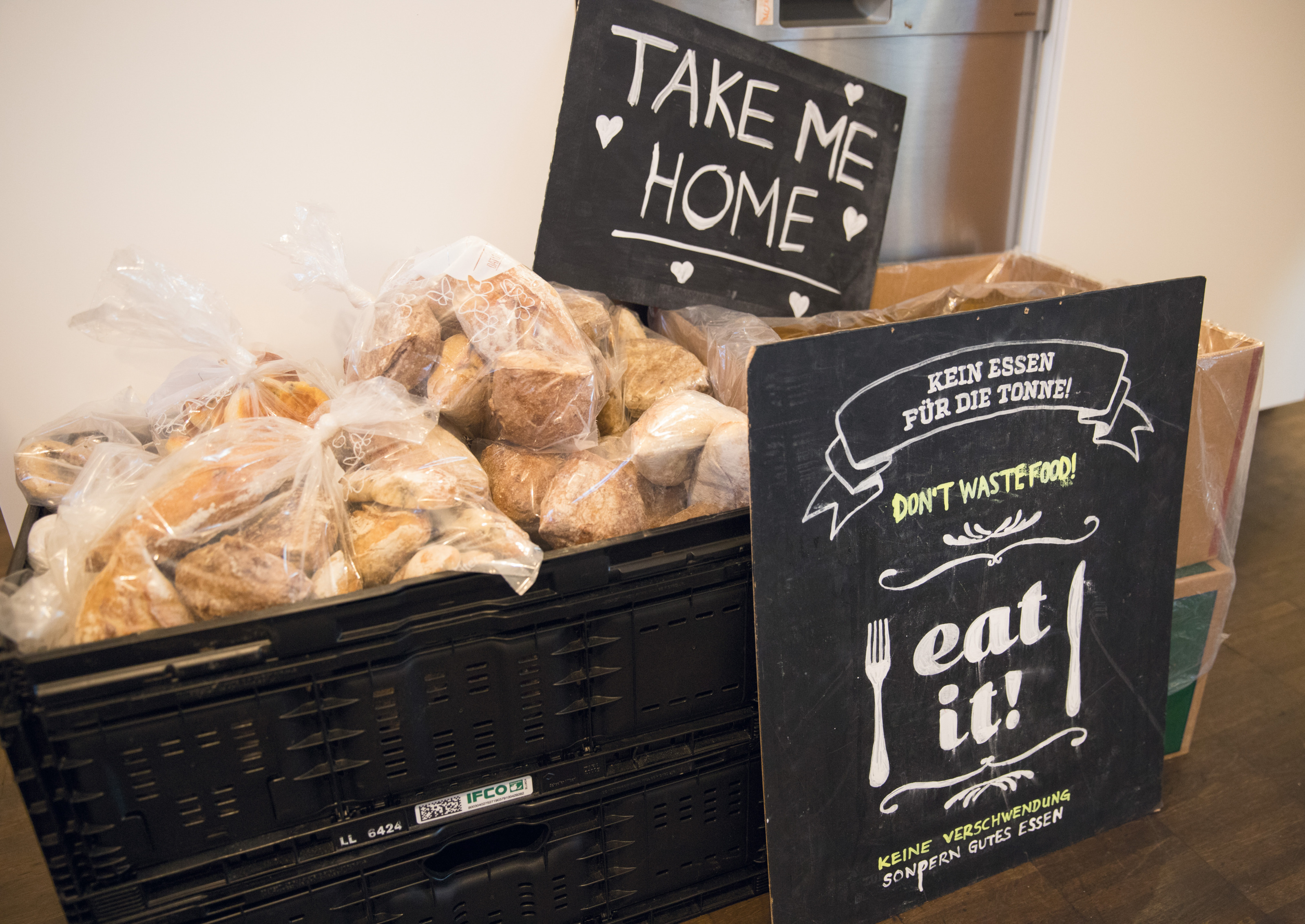 Kisten mit abgepackten Lebensmitteln, darauf und davor Tafeln lehnend mit Schriftzug Take me Home und Kein Essen f&uuml;r die Tonne, don't waste food