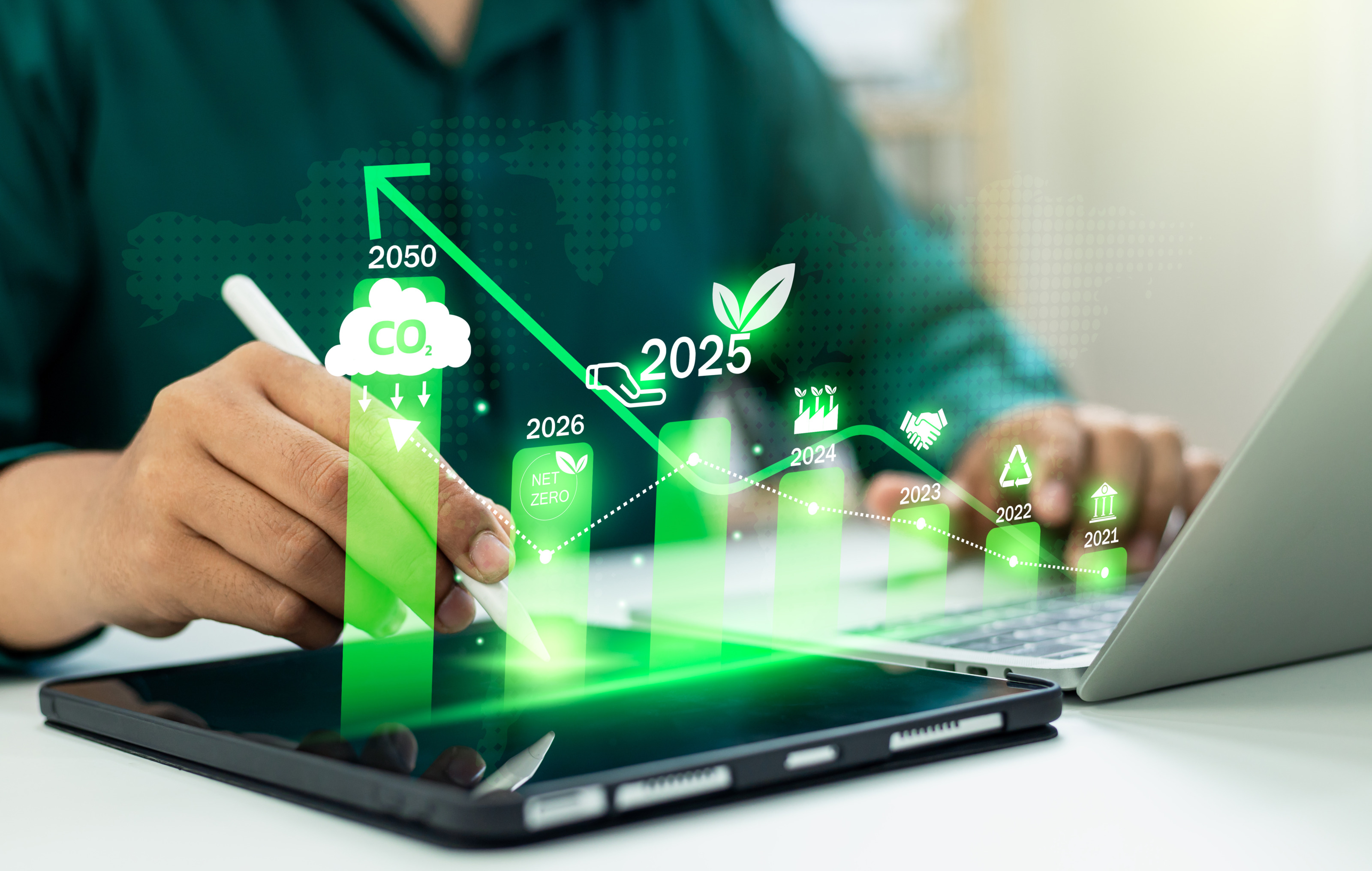 Nahaufnahme einer Person, die auf einem Laptop tippt, sie hat einen Stift in der Hand, daneben liegt ein Tablet. Man sieht ein gr&uuml;nes Balkendiagramm mit den Zahlen 2025 und 2025 und Symbolen: Wolke mit Co2, Bl&auml;tter, Kreislauf, Parlament, H&auml;nde.  