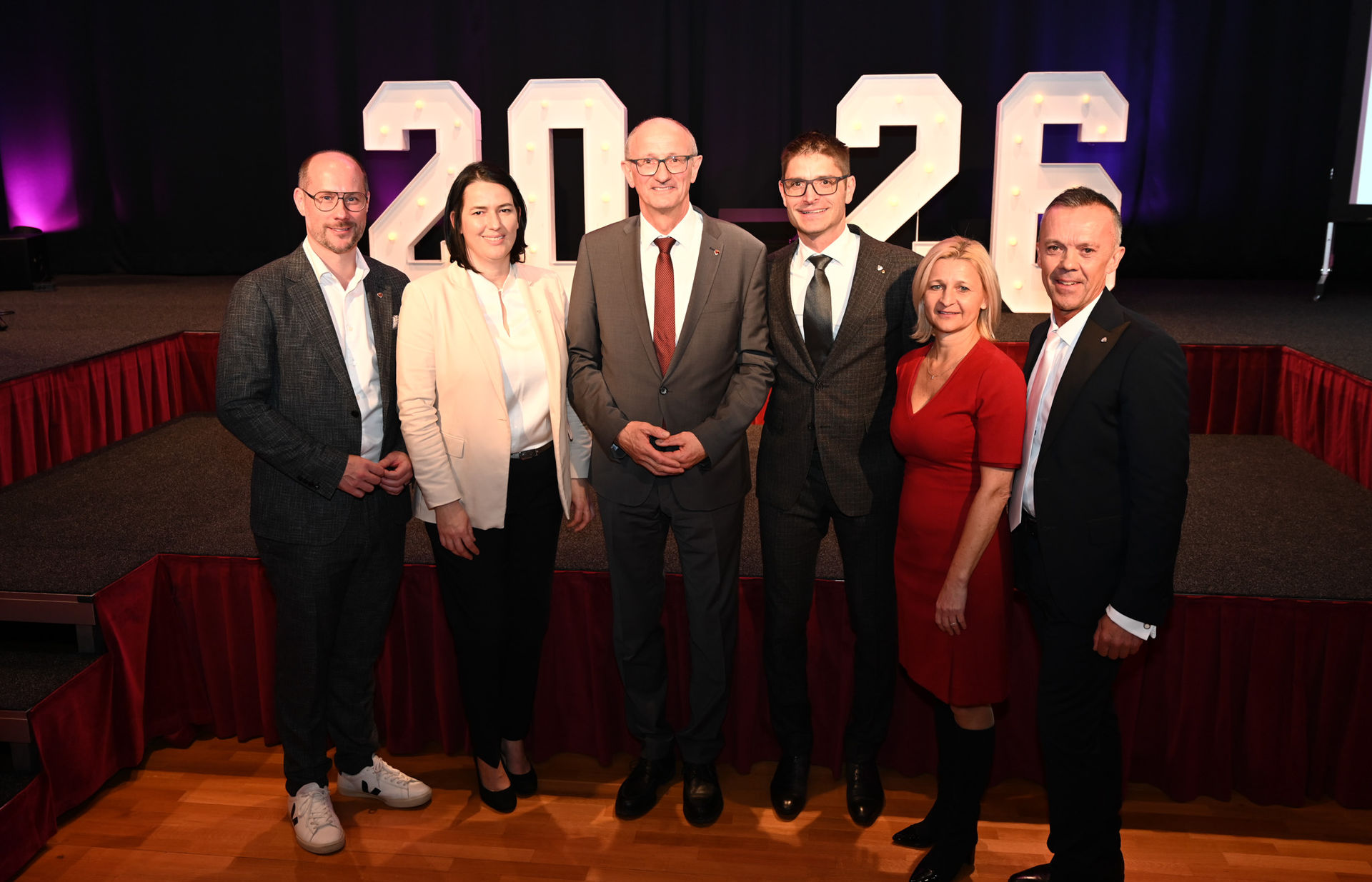 Sieben Personen stehen nebeneinander vor einer B&uuml;hne und blicken in die Kamera. Im Hintergrund sind gro&szlig;e, beleuchtete Zahlen &bdquo;2026&ldquo; zu sehen. Die Personen tragen formelle oder halbformelle Kleidung. Die Person in der Mitte steht leicht vor den anderen, alle stehen eng beieinander. Der Raum ist ein Veranstaltungssaal mit rotem B&uuml;hnenvorhang und Holzboden. Die Stimmung wirkt offiziell und festlich.