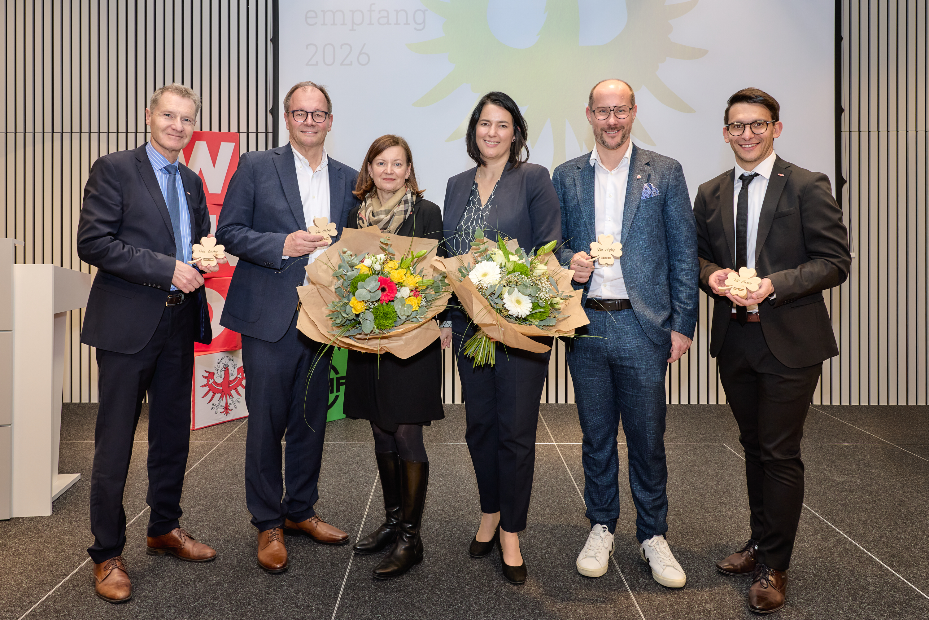 Sieben Personen stehen nebeneinander auf einer B&uuml;hne und blicken in die Kamera. Zwei Personen in der Mitte halten Blumenstr&auml;u&szlig;e, die anderen jeweils eine kleine Holzfigur in der Hand. Alle Personen tragen formelle oder halbformelle Kleidung. Im Hintergrund ist eine Projektionsfl&auml;che mit einem Adler-Symbol und der Aufschrift &bdquo;empfang 2026&ldquo; zu sehen. Links steht ein rotes Logo-Element, das zu einer Organisation geh&ouml;rt. Der Raum wirkt wie ein Veranstaltungs- oder Festsaal.