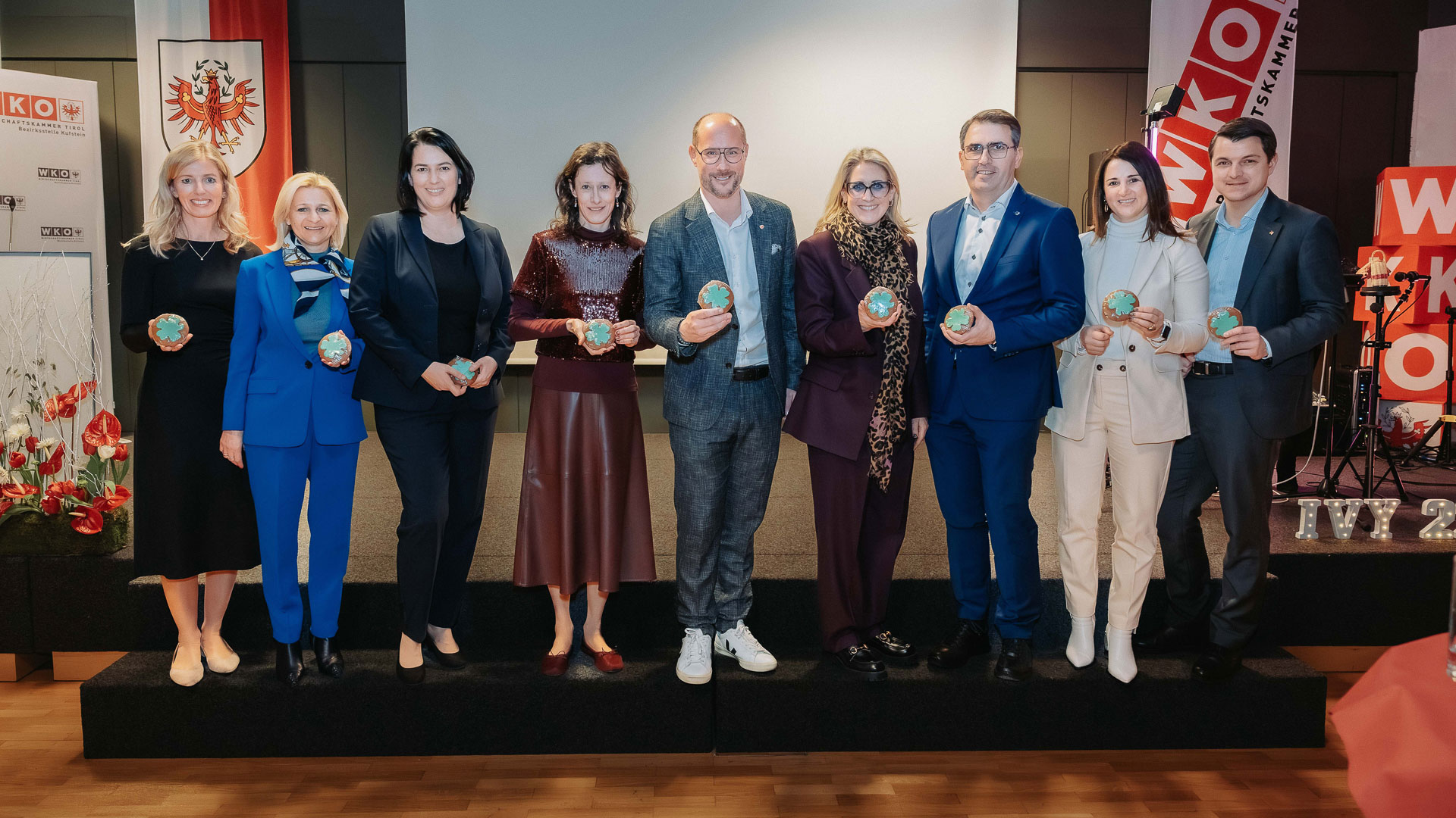 Zehn Personen stehen nebeneinander auf einer B&uuml;hne und halten jeweils ein rundes, dekoratives Objekt in der Hand. Alle Personen sind elegant gekleidet. Im Hintergrund sind Banner der Wirtschaftskammer Tirol sowie eine Tiroler Fahne zu sehen. Die B&uuml;hne ist beleuchtet, der Raum wirkt wie ein offizieller Veranstaltungs- oder Festsaal. Vor der B&uuml;hne ist ein Holzboden sichtbar, rechts stehen technische Ger&auml;te f&uuml;r Ton oder Licht.