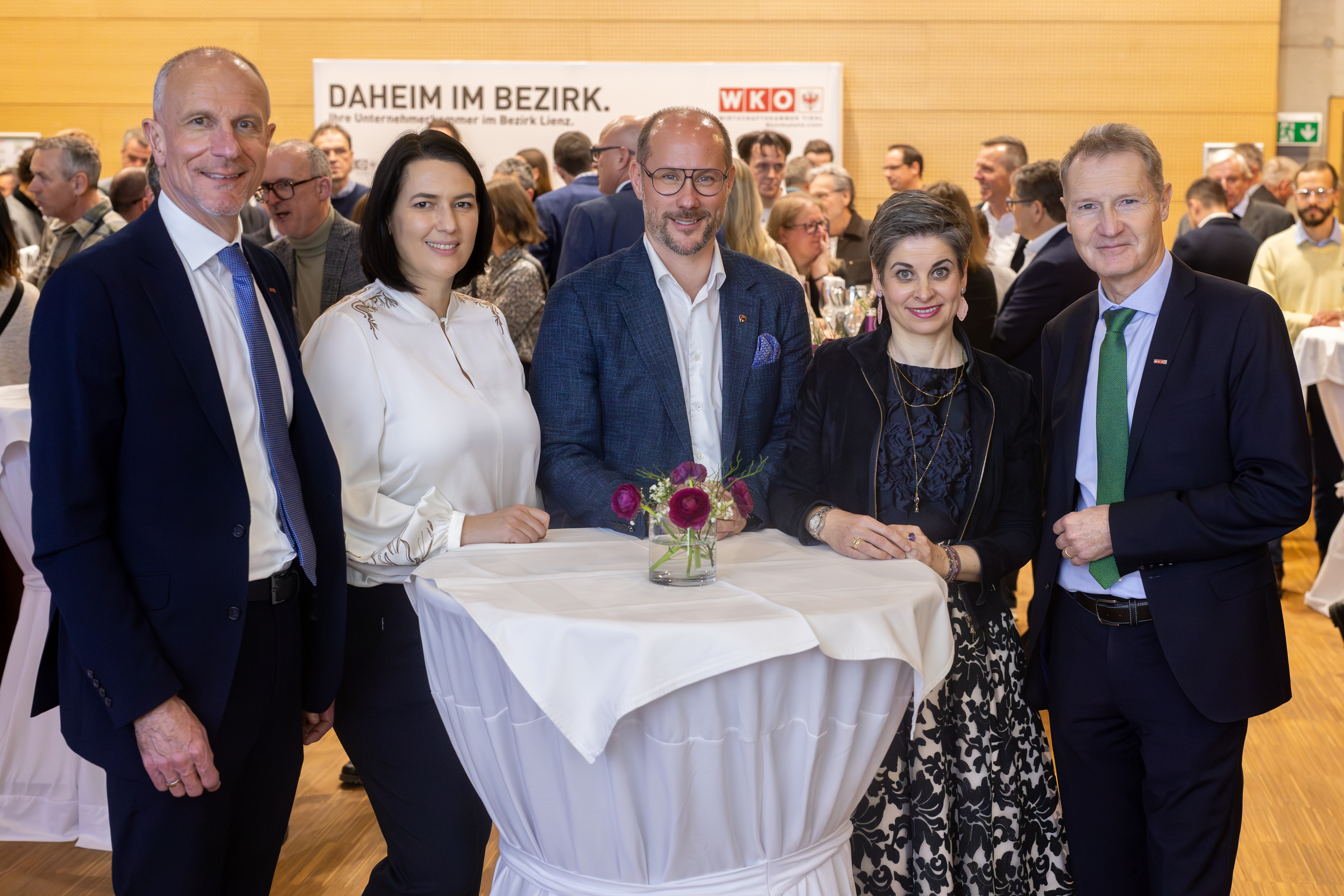 F&uuml;nf Personen stehen gemeinsam an einem Stehtisch in einem Veranstaltungsraum. Alle sind elegant gekleidet und blicken in die Kamera. Auf dem Tisch steht eine kleine Blumenvase. Im Hintergrund sind viele weitere Personen zu sehen, die sich unterhalten. Hinter der Gruppe h&auml;ngt ein Banner mit der Aufschrift &bdquo;Daheim im Bezirk&ldquo; und dem Logo der Wirtschaftskammer.