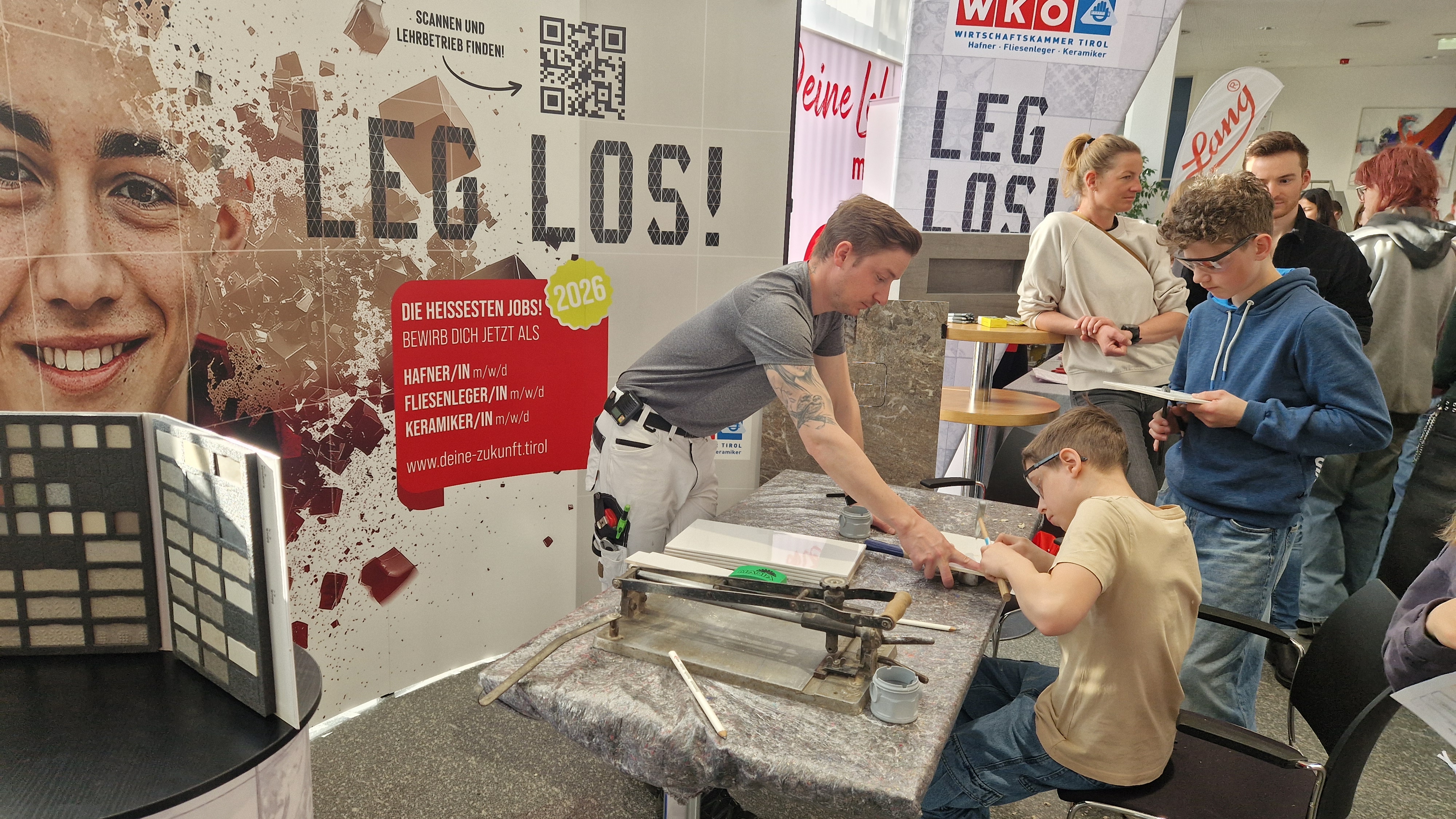 Ein Arbeiter zeigt einem Jugendlichen das Fliesenlegen an einem Messestand der Wirtschaftskammer Tirol. Auf dem Tisch liegen Fliesen, Werkzeuge und ein Fliesenschneider. Im Hintergrund wirbt ein gro&szlig;es Plakat f&uuml;r Lehrberufe wie Hafner:in, Fliesenleger:in und Keramiker:in.