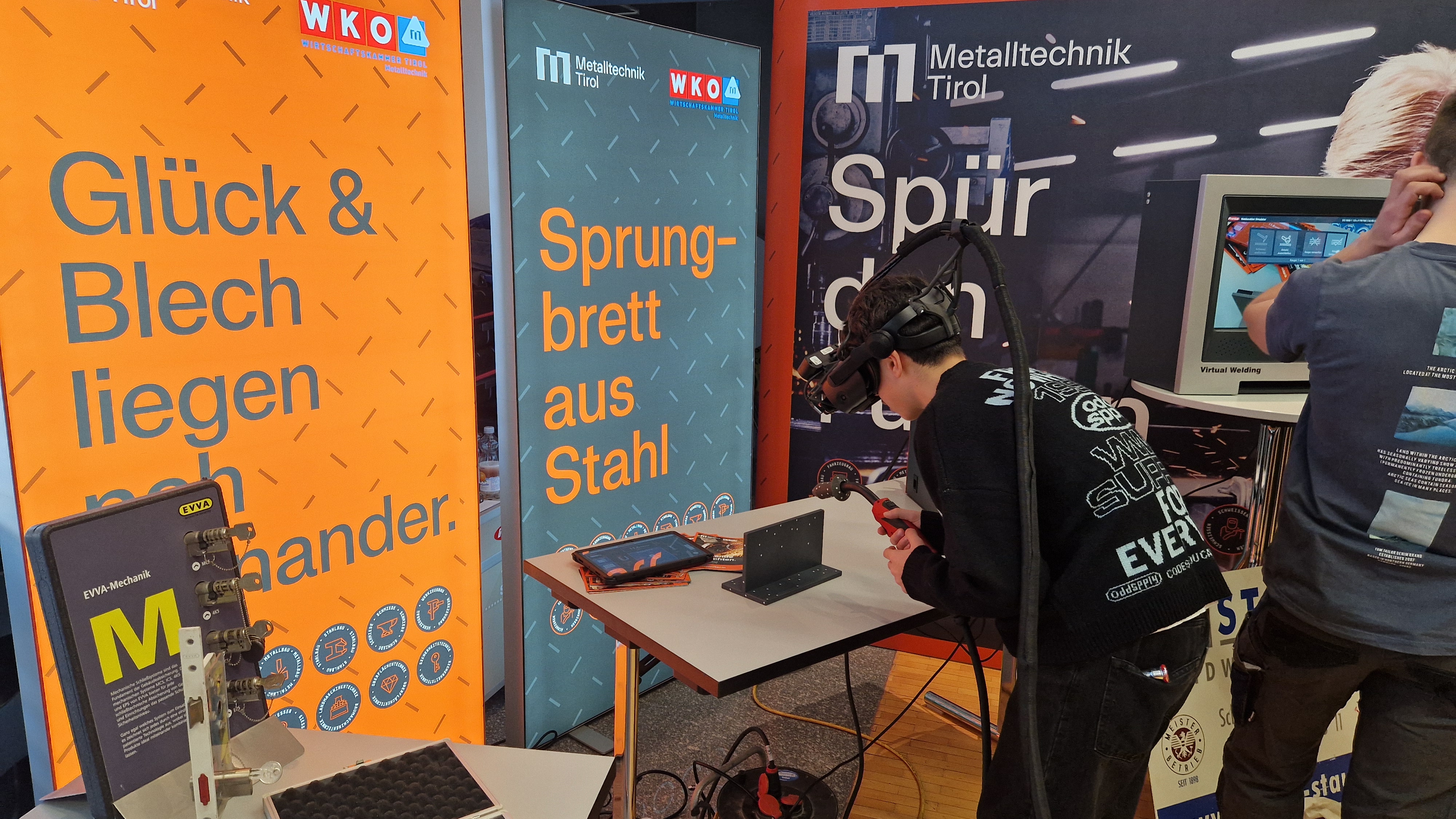 Ein Jugendlicher testet Virtual-Reality-Schwei&szlig;en am Stand von Metalltechnik Tirol. Mit VR-Brille und Schwei&szlig;ger&auml;t simuliert er eine Schwei&szlig;arbeit, w&auml;hrend ein Bildschirm die Anwendung zeigt. Infotafeln im Hintergrund erkl&auml;ren die Ausbildung im Metallbereich.