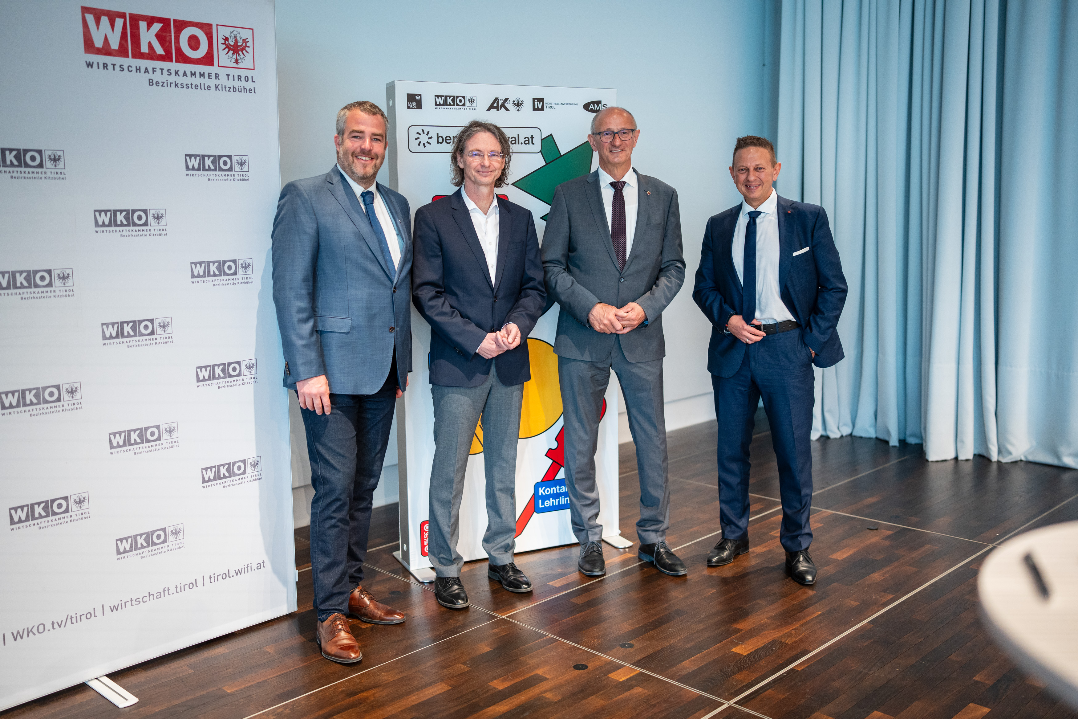 Vier M&auml;nner in Anz&uuml;gen stehen nebeneinander in einem Veranstaltungsraum der Wirtschaftskammer Tirol, WK-Bezirksstelle Kitzb&uuml;hel. Sie blicken in die Kamera und l&auml;cheln. Im Hintergrund sind WKO-Roll-ups und ein Informationsstand zu sehen. Der Raum ist hell beleuchtet, der Boden aus Holz.