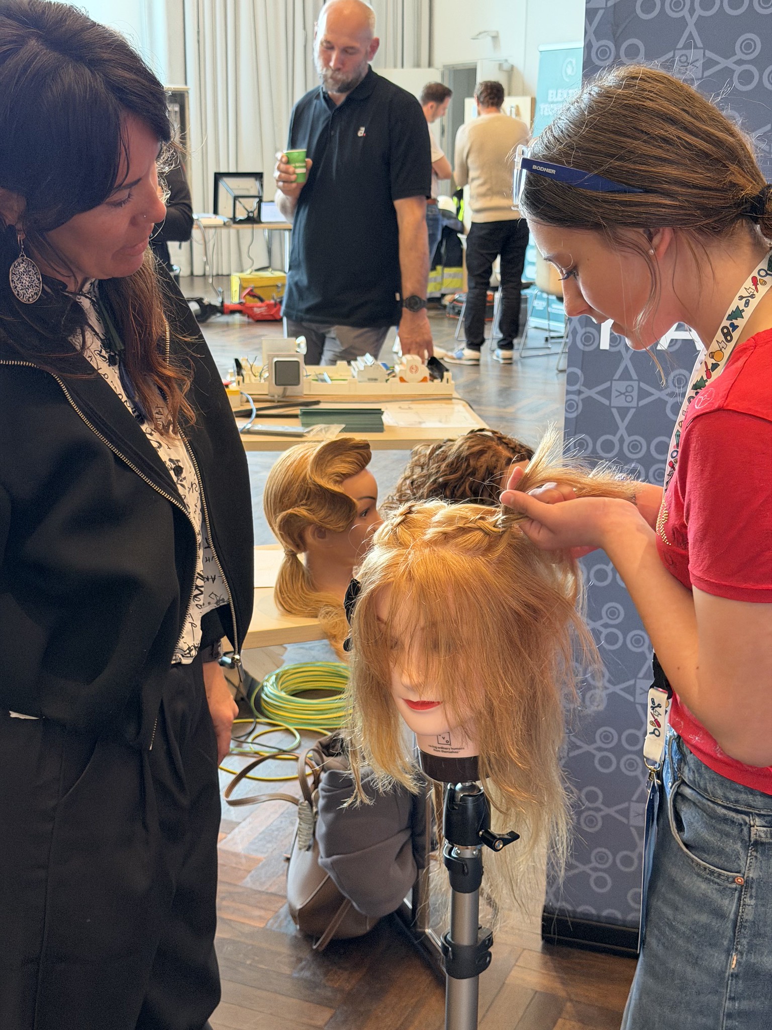 Eine junge Frau flechtet konzentriert die Haare einer &Uuml;bungspuppe, w&auml;hrend eine zweite Frau danebensteht und zusieht. Im Hintergrund sind weitere Personen und Arbeitsstationen zu sehen. Die Szene wirkt wie ein Praxis-Workshop im Friseurhandwerk.