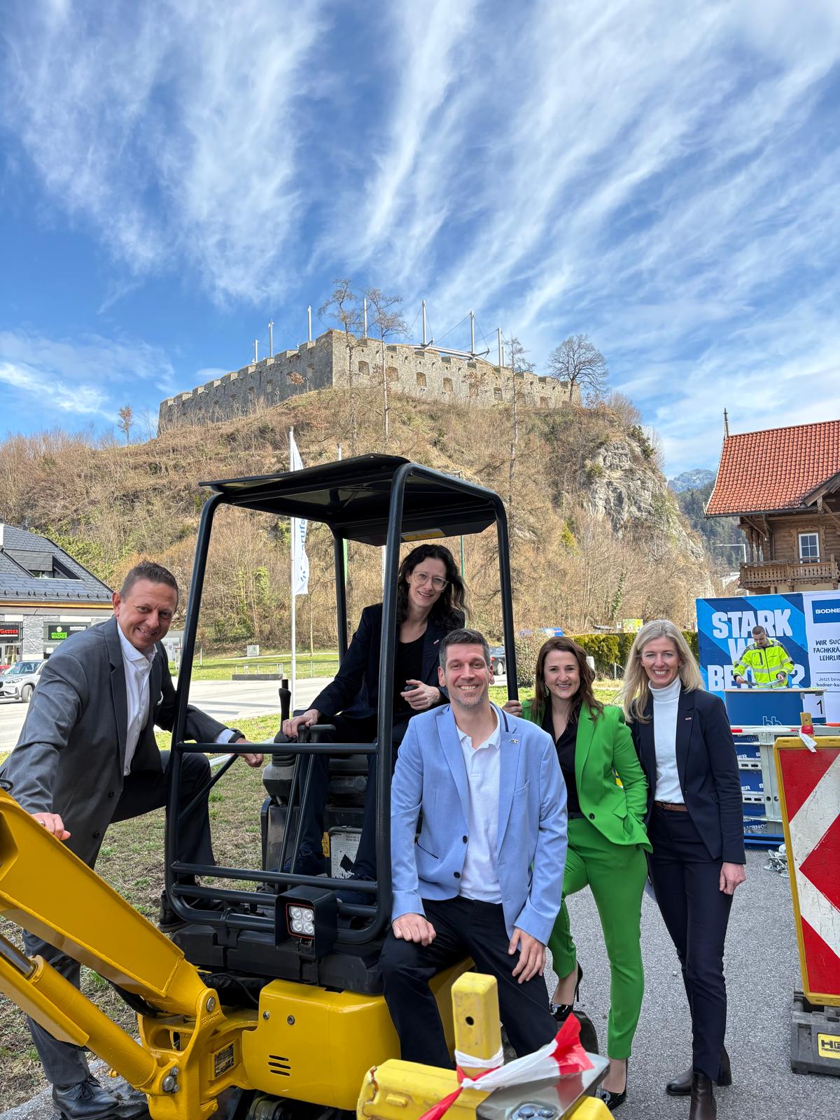 Gruppe von f&uuml;nf Personen steht und sitzt auf einem Minibagger im Freien, im Hintergrund eine Festungsanlage; aufgenommen beim Berufsfestival Kufstein.